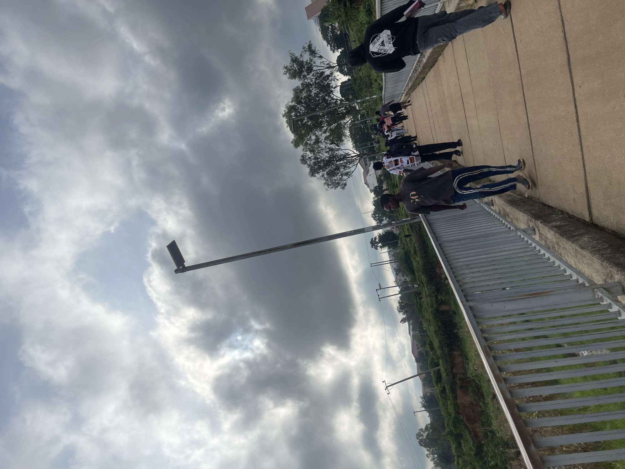 The image depicts an elevated pedestrian walkway with a metal railing on the left side, leading into the distance. Several individuals are walking on the walkway. The sky is overcast with dramatic, dark clouds, partially obscuring the sun. In the background, there are trees, some buildings, and utility poles with power lines, suggesting an urban or semi-urban environment. The walkway appears to be made of concrete. The overall scene suggests a daytime, possibly pre-rainy weather condition. This location is identified as Rafin Par, Nigeria.