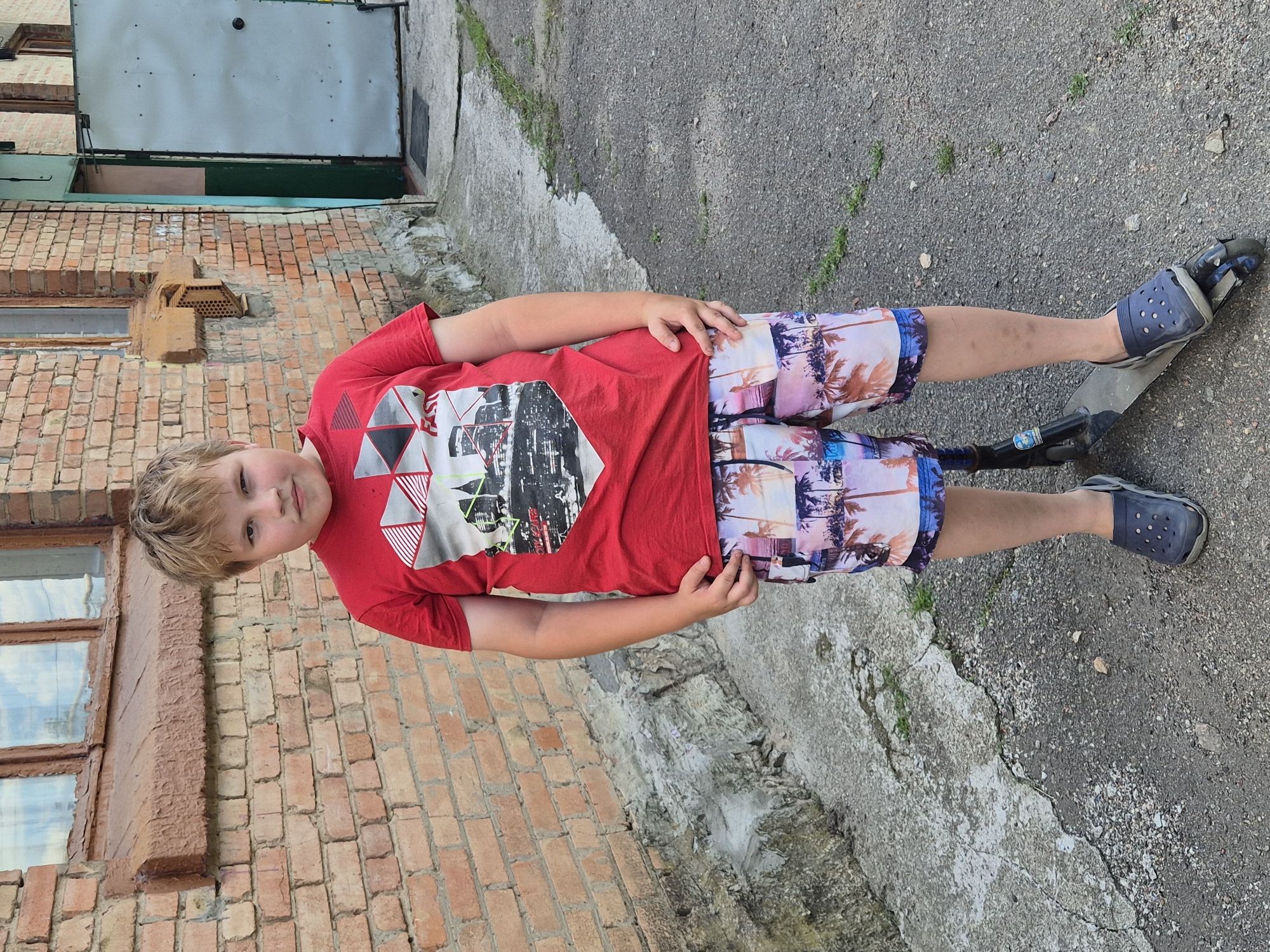 A young boy with blond hair, wearing a red t-shirt and colorful shorts with a palm tree print, stands on a scooter. He is looking at the camera with a slight smile. The background consists of a brick building with windows, a concrete surface, and some green vegetation. The overall scene suggests an outdoor setting, possibly a yard or alleyway, during the daytime. The boy's attire and the scooter indicate a casual, recreational activity. The image is oriented vertically, with the boy positioned in the center. There is no visible text in the image.
