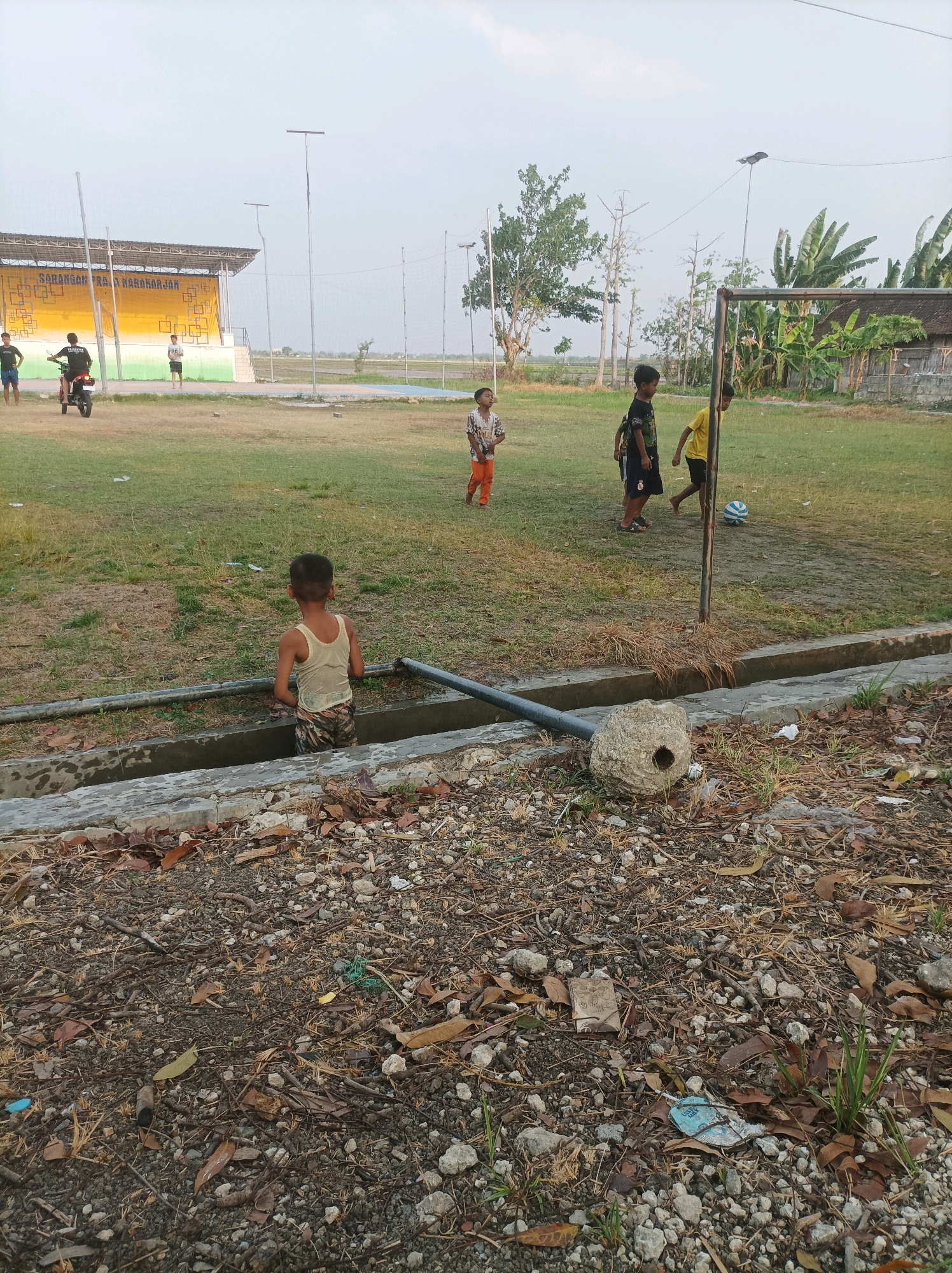 In Sarangan, Indonesia, children are playing soccer in a grassy field with a soccer goal. One boy in an orange shirt and white shorts runs towards the ball, while another in a yellow shirt stands near the goal. Two other boys are visible further in the background, one riding a motorcycle. The field is bordered by a concrete structure and a ditch. The sky is overcast, and the overall lighting suggests it is late afternoon.