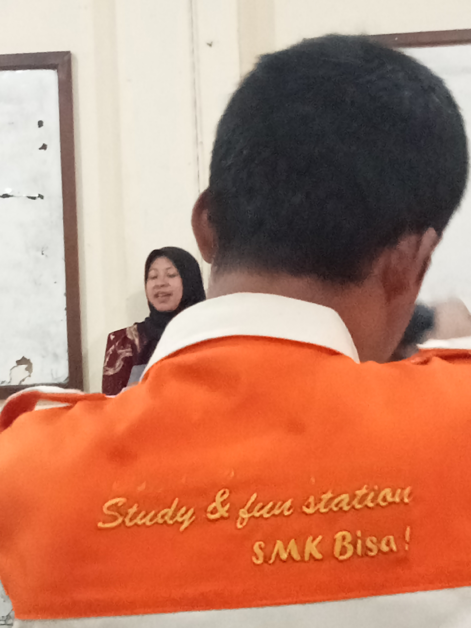 The image shows a close-up view of the back of a person wearing an orange shirt with a white collar. Embroidered on the shirt in yellow is the text "Study & fun station" on the top line and "SMK Bisa!" on the bottom line. In the background, a woman wearing a black hijab and a patterned maroon top is visible. She is looking to her left and appears to be speaking. To the far left, a portion of a whiteboard with some marks on it is visible, framed by a brown border. The environment appears to be indoors, possibly a classroom or a meeting room. The location is identified as Secang, Indonesia.