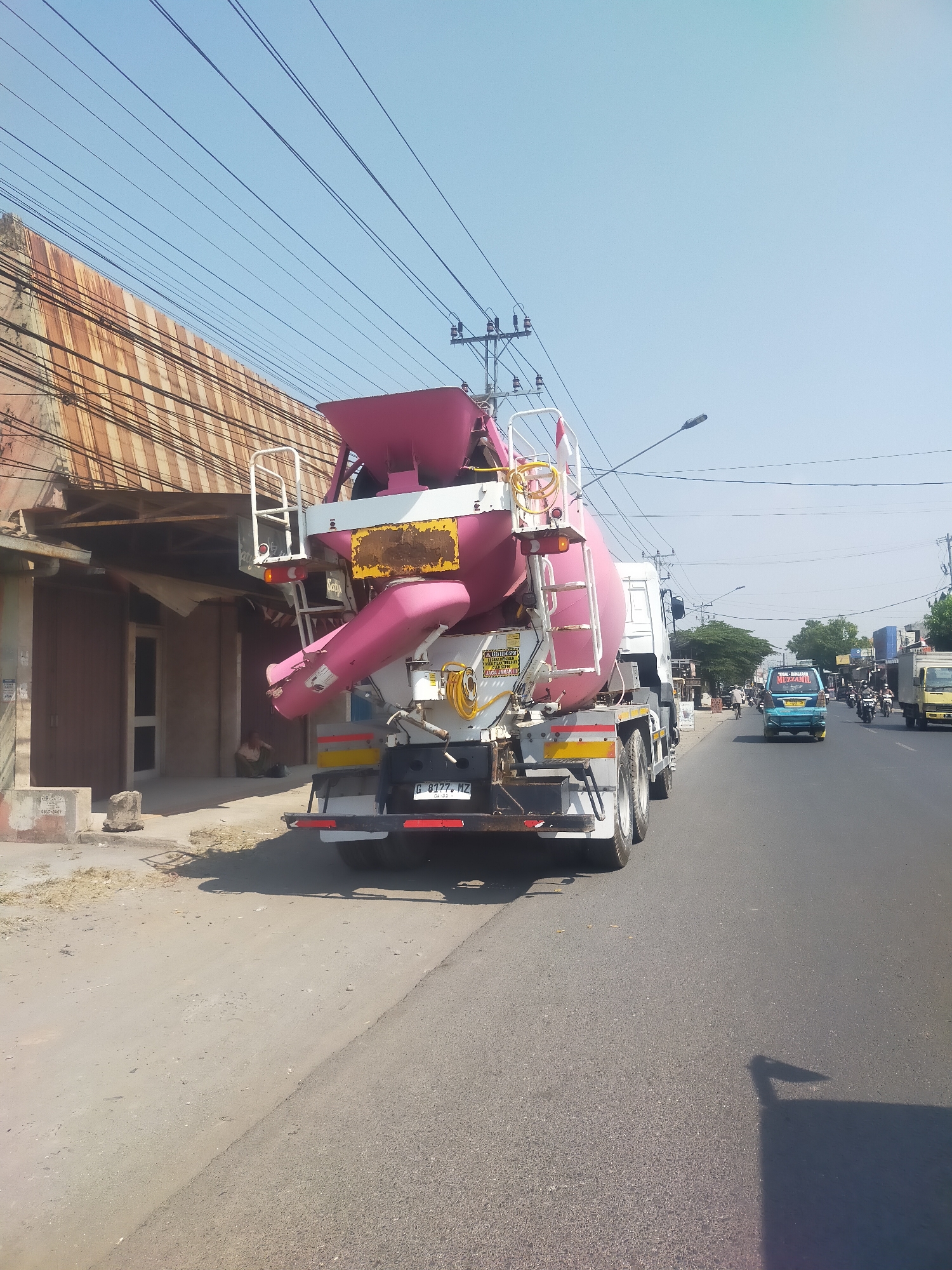 A concrete mixer truck, primarily pink and white with some black, is parked on the right side of a paved road in Badiran, Indonesia. The truck has a large cylindrical drum that is tilted forward. A white ladder is attached to the side of the drum. The truck's license plate reads "G 8177 MZ". In the background, there is a building with a corrugated metal roof and brown doors, and a person is sitting on the ground in front of the building. Several power lines crisscross the sky. Other vehicles, including a blue van and a yellow truck, are visible on the road ahead. The scene appears to be during the daytime with a clear blue sky.