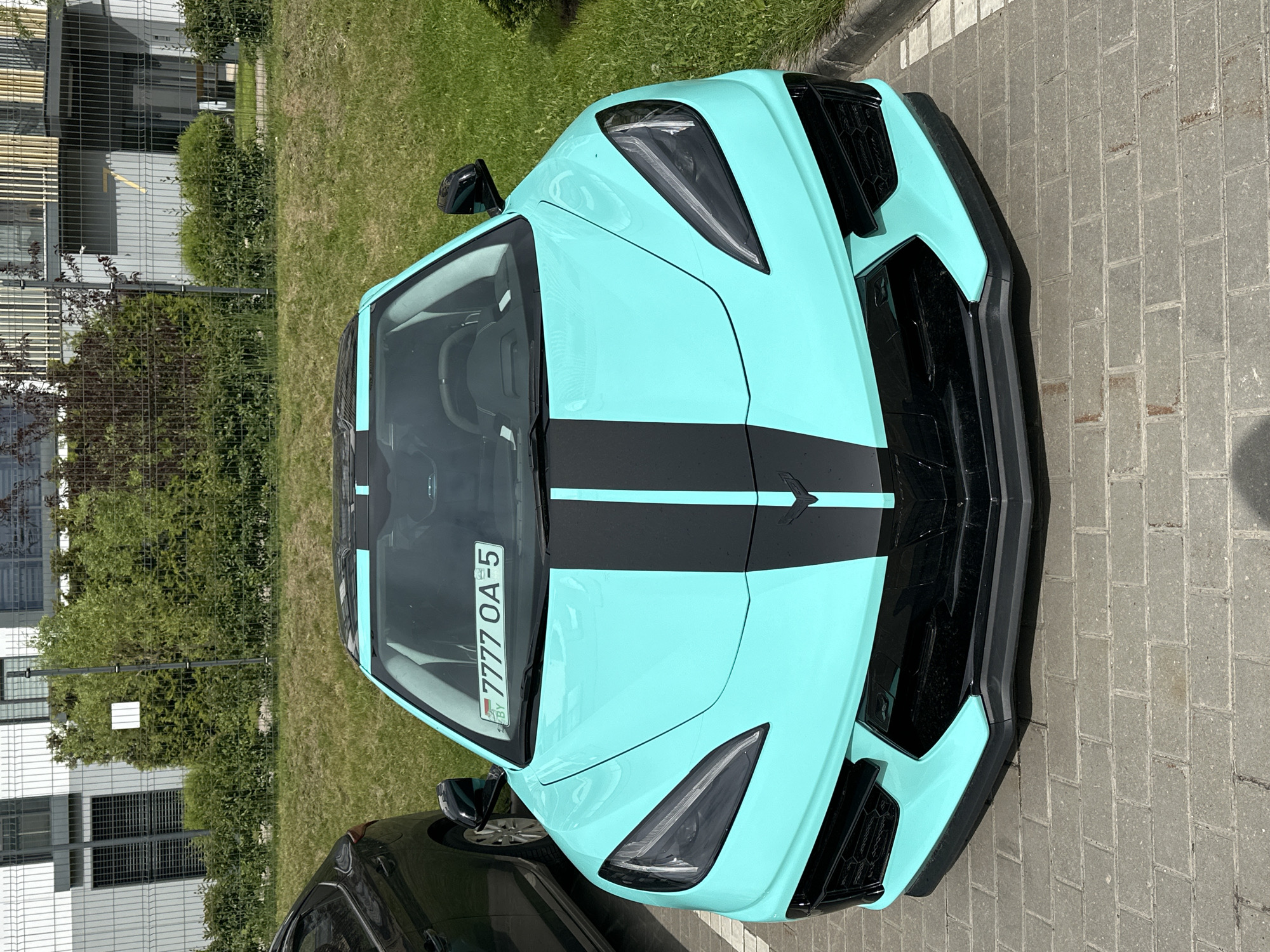 The image shows the front of a turquoise Chevrolet Corvette with black stripes parked in Бараўлянскі сельскі Савет, Belarus. The license plate "7777 OA-5" is visible. The car is on a paved area next to a grassy area with bushes and a building in the background. Another dark-colored car is partially visible to the lower left of the Corvette. The weather appears to be sunny.