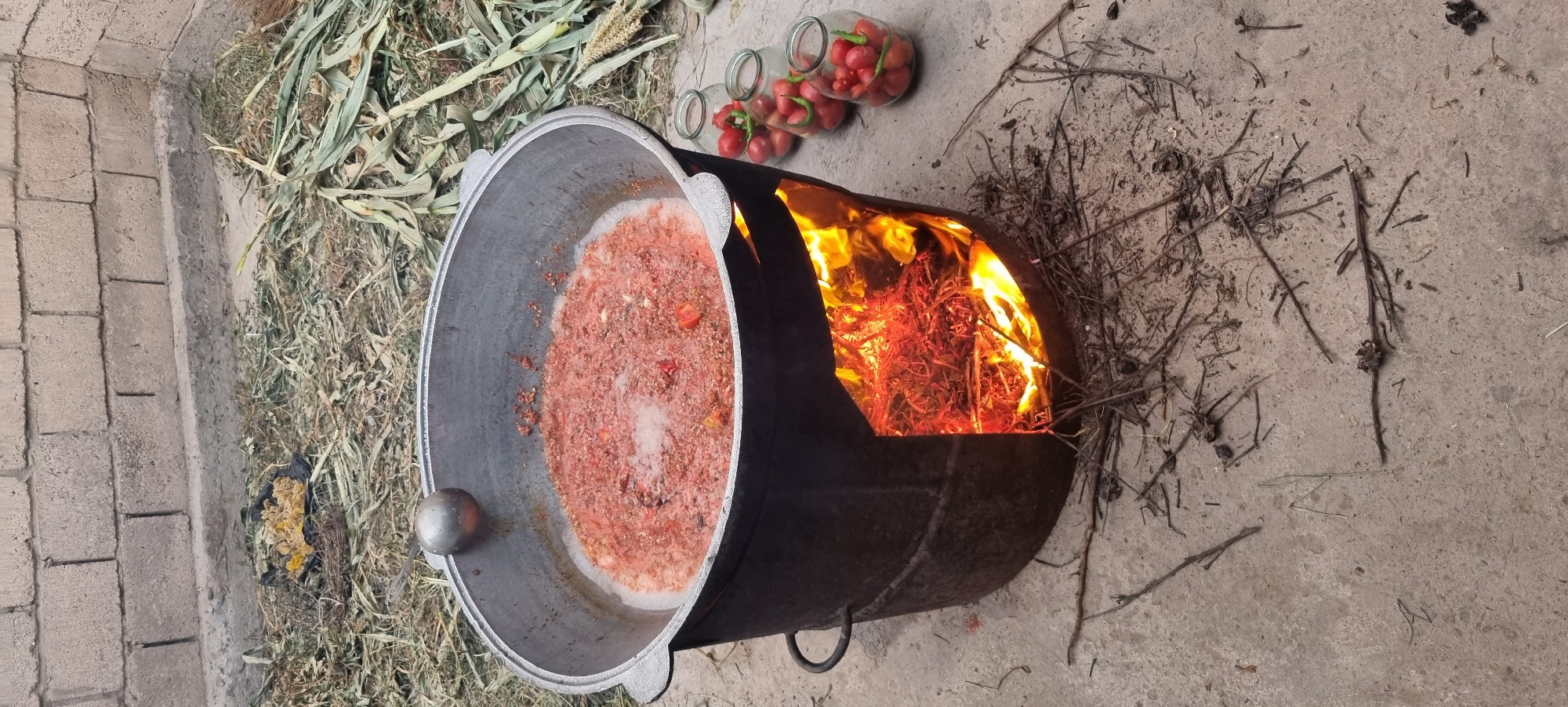 A large cast-iron cauldron filled with a simmering red mixture, likely tomatoes and spices, is heated by a fire burning in an open hearth beneath it. The cauldron is situated on a concrete surface in an outdoor setting. To the right of the cauldron, three glass jars filled with small red tomatoes and green chili peppers are visible. In the background, a rough brick wall stands behind a patch of dried grass and corn stalks. The overall scene suggests food preparation, possibly canning or cooking a traditional dish. The location is identified as Jomi, Tajikistan.
