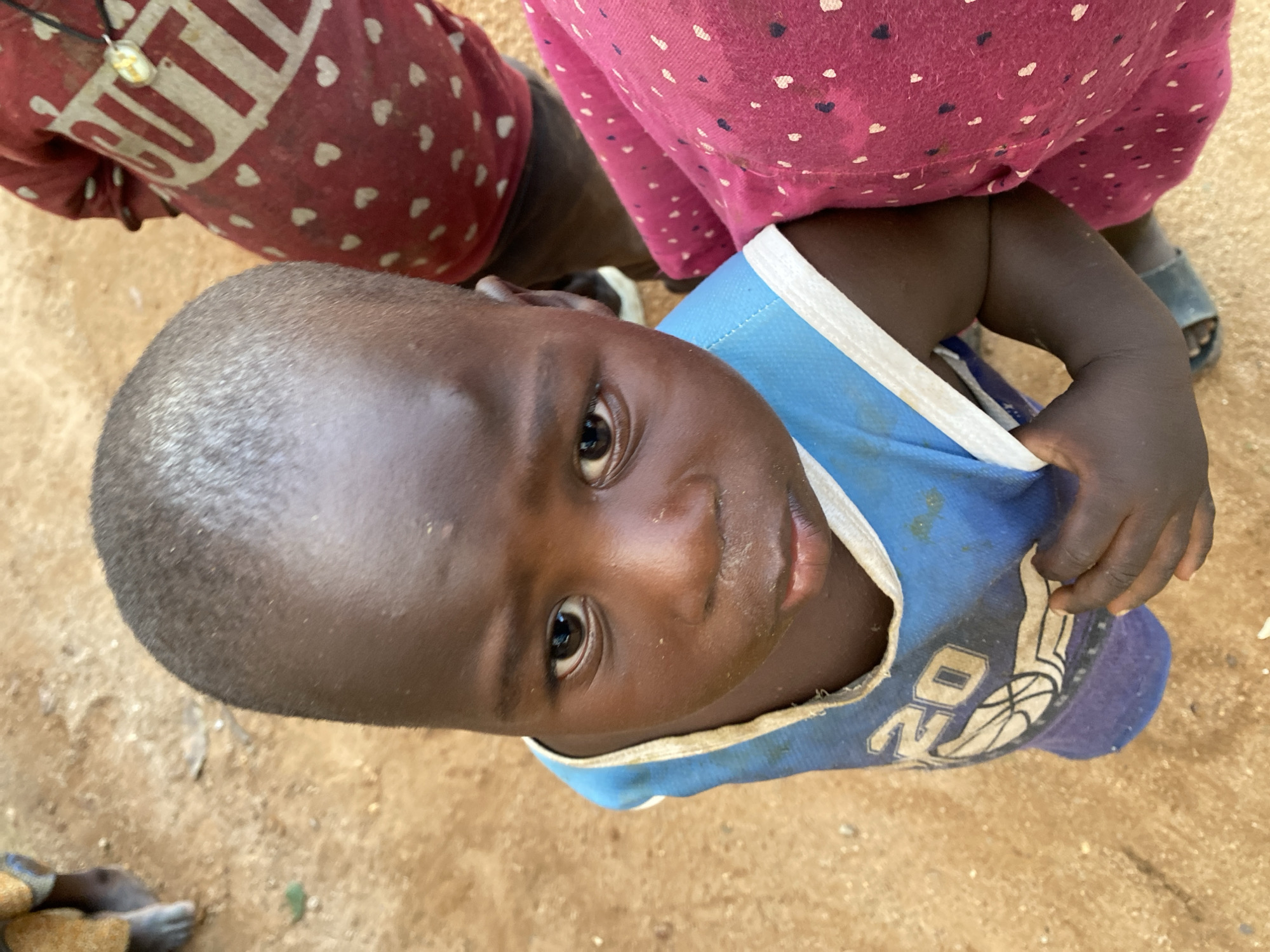 A close-up, top-down shot shows a young Black child in Riyom, Nigeria. The child gazes upwards at the camera with a solemn expression. The child wears a blue and white tank top with a basketball-themed graphic. Partial views of two adults are visible: one wears a red shirt with white hearts and the word "CUTIE" printed on it; the other wears a pink dress with small white dots. The ground appears to be sandy or dirt-covered.