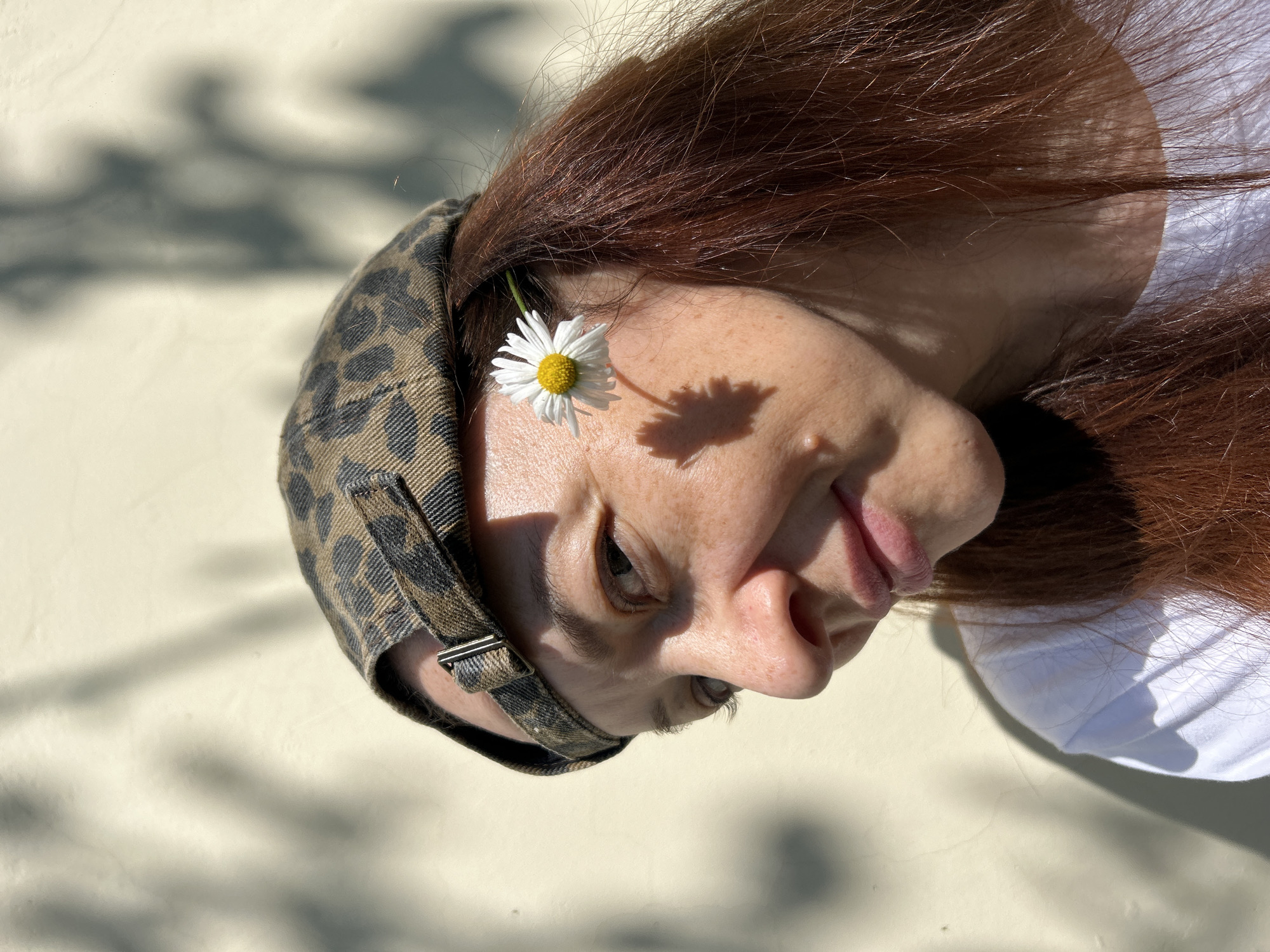A medium shot shows a woman with reddish-brown hair wearing a camouflage baseball cap and a white t-shirt. She is outdoors with a plain, light-colored wall behind her, on which shadows of foliage are cast. A small white daisy with a yellow center is tucked behind her ear. The woman is looking to her left, and her face is partially illuminated by sunlight, creating a shadow of the daisy on her cheek. The woman has freckles and a mole on her cheek. The image captures a serene moment in an outdoor urban setting, possibly a park or a residential area. There is no discernible information within the image to indicate the specific location in Chaikovsky, Russia.