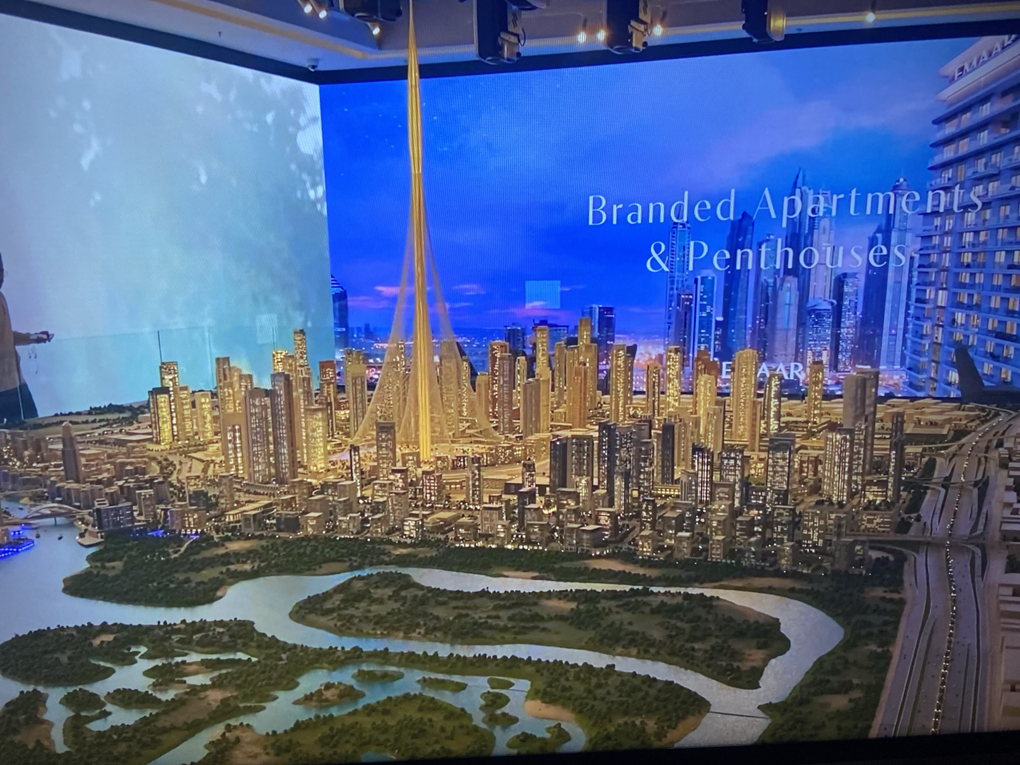 The image shows a scale model of a city, likely Dubai, featuring numerous high-rise buildings, a river with islands, and a highway. The tallest structure is a tower with a cable-supported design. A large screen in the background displays a digital image of the city skyline, with the text "Branded Apartments & Penthouses" and the logo "EMAAR". The model is illuminated, highlighting the architectural details. A person is partially visible on the left edge of the image. The scene appears to be an exhibit or display showcasing the city's architecture and real estate developments.