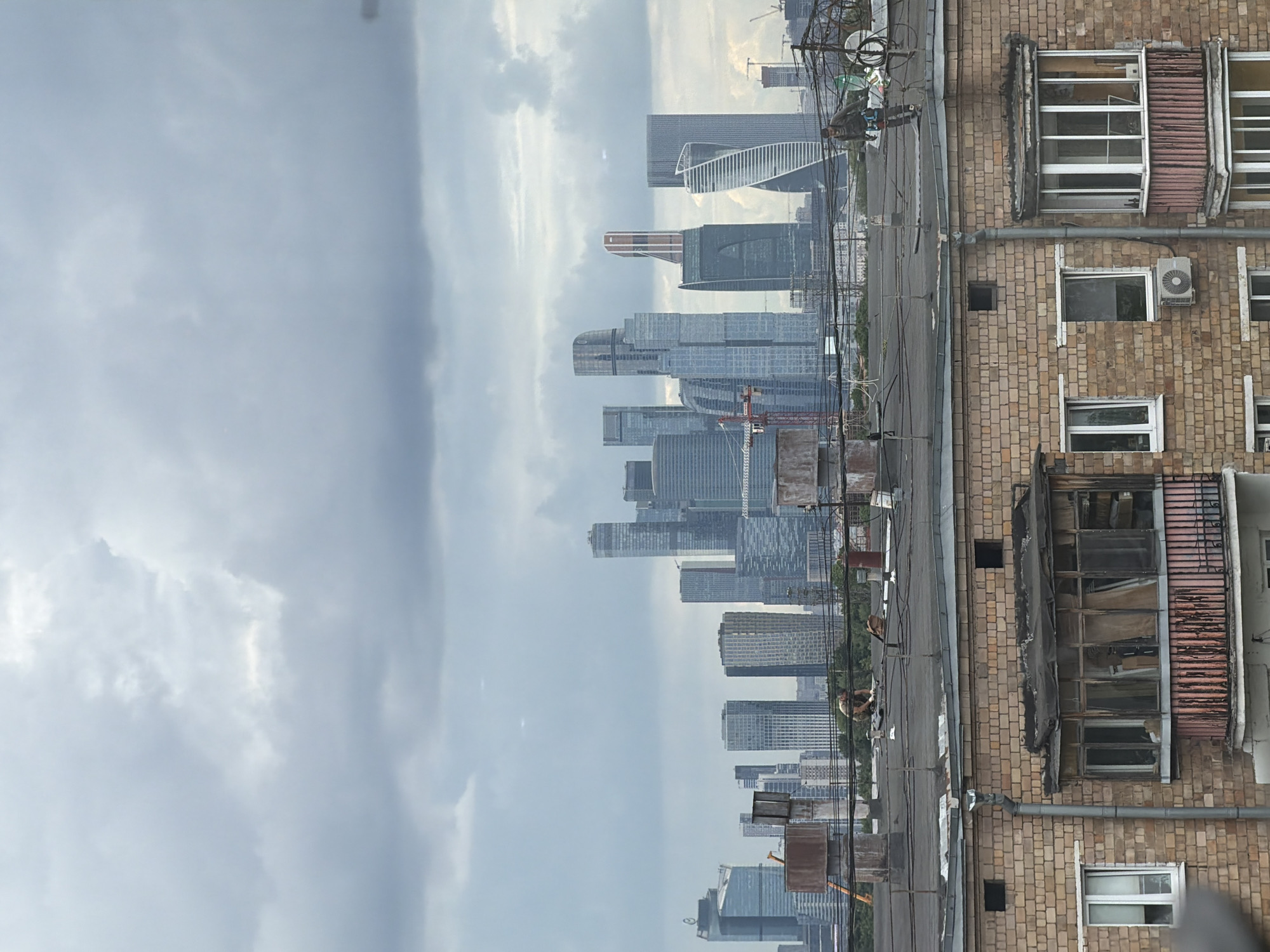 The image shows a wide shot of a city skyline under a cloudy sky. In the foreground, the flat roof of a brick apartment building is visible, with several ventilation pipes and a satellite dish. Two figures are on the roof, one appearing to be working on something. A second, older brick building with a balcony is partially visible on the lower left. In the background, a dense cluster of modern skyscrapers dominates the horizon, with a construction crane visible among them. The sky is overcast, with large, grey clouds. The overall impression is an urban scene with a stark contrast between older and newer architecture. The location is unknown.