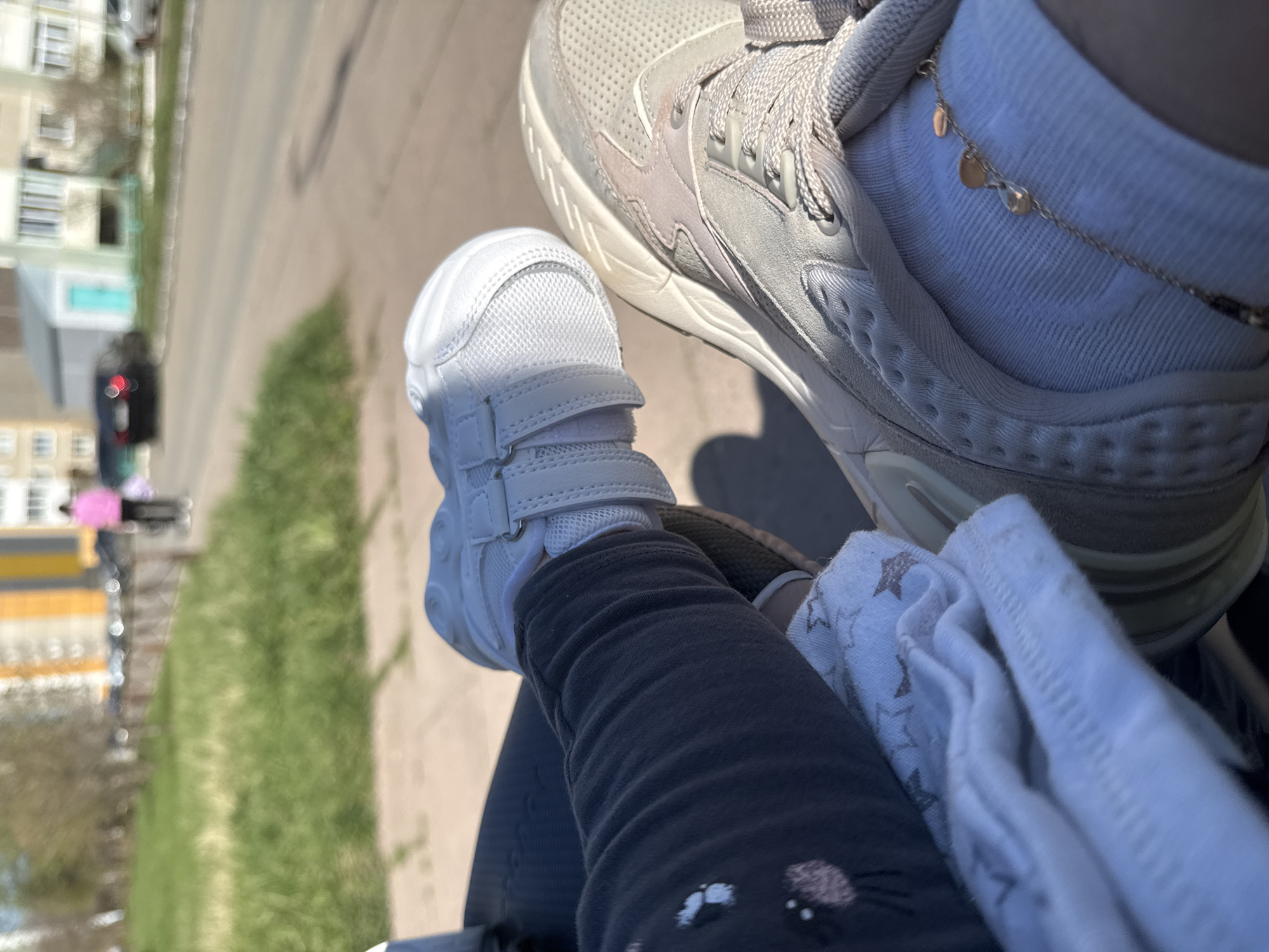 The image, taken from an elevated perspective, shows a portion of a city, possibly Chaikovsky, Russia. In the foreground, a baby's white shoe and dark pants with a white blanket are visible. Next to the baby's shoe is an adult's grey sneaker, worn with a white sock that features a gold-colored anklet. Outside, the grass and the sidewalks are visible. Beyond that are city buildings. There are pedestrians outside, including one person carrying a pink umbrella.