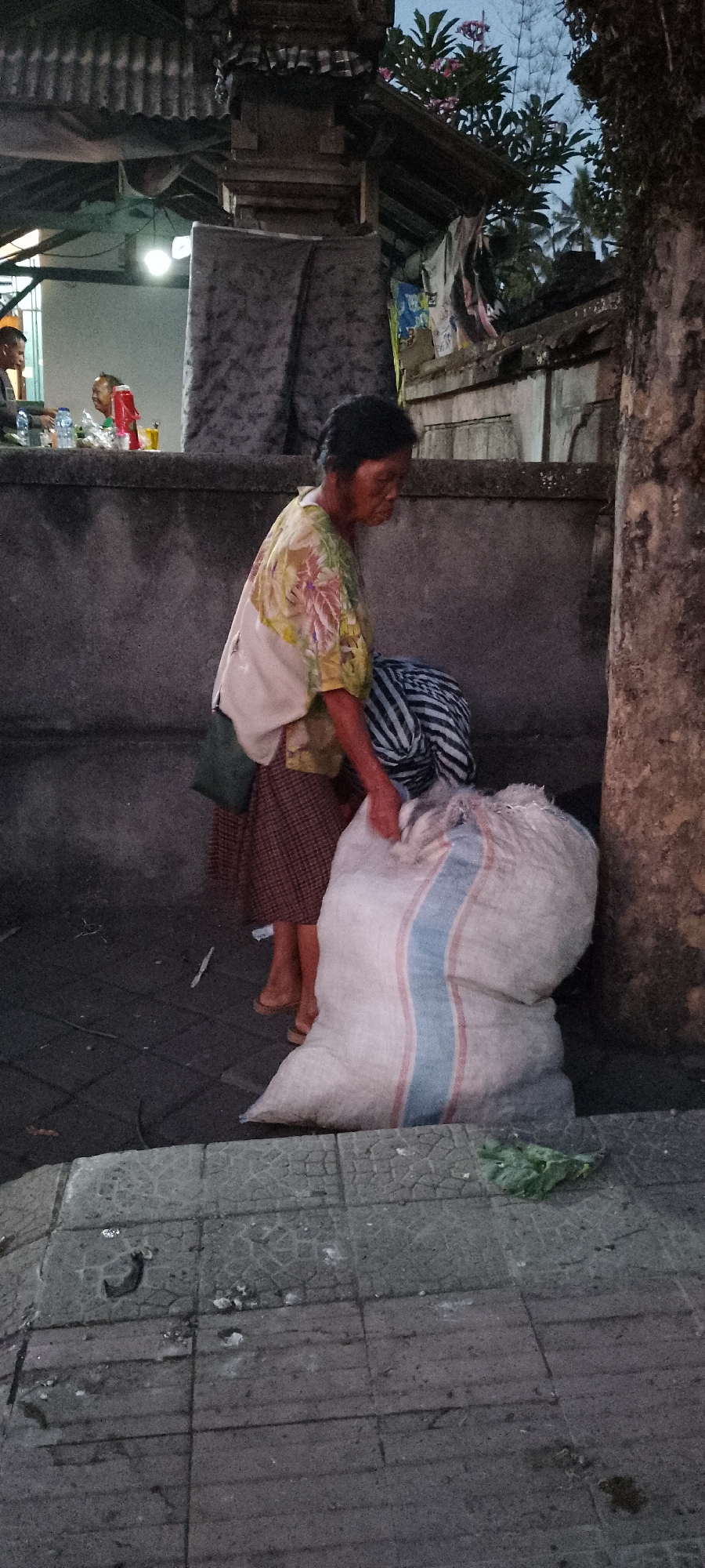 An older Asian woman with dark hair is standing near a large, white burlap sack that is tied at the top and has two vertical stripes of blue and red. She is wearing a light-colored, short-sleeved shirt with a floral print in shades of yellow, green, and pink, and a brown and white checkered skirt. A dark green bag is slung over her left shoulder. She is reaching down with her right hand towards the sack. Behind her, a dark-gray concrete wall is visible. To the left of the woman, there appears to be a covered outdoor area with a metal roof and some lighting. In the background of this covered area, several people are seated at tables, and there are bottles and red and yellow containers on the tables. The scene is outdoors, with trees and foliage visible in the upper right background. The ground in the foreground is paved with gray square tiles, some of which are cracked, and there are scattered debris. This scene is located in Pekutatan, Indonesia.