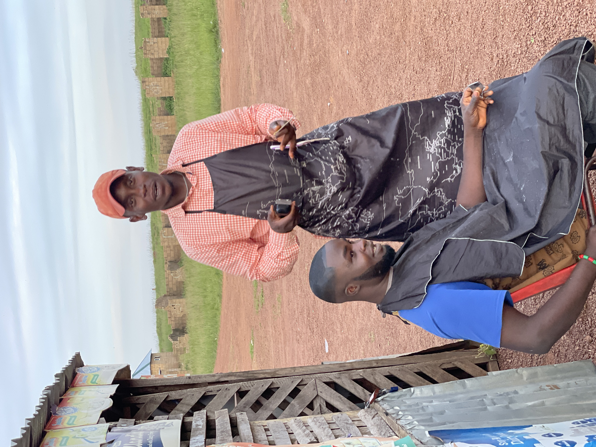 In Durumi, Nigeria, a barber stands behind a seated customer. The barber, wearing an orange baseball cap and a red and white checkered shirt under a black apron adorned with a world map pattern, holds a clippers in his left hand and a comb in his right. The customer, dressed in a blue t-shirt and a black barber cape, is seated and looking to his right. To the left of the frame, a makeshift shelter constructed from corrugated metal and wooden planks displays several posters. In the background, unfinished brick structures and a grassy field extend towards the horizon under a cloudy sky. The ground is a dusty, reddish-brown earth.