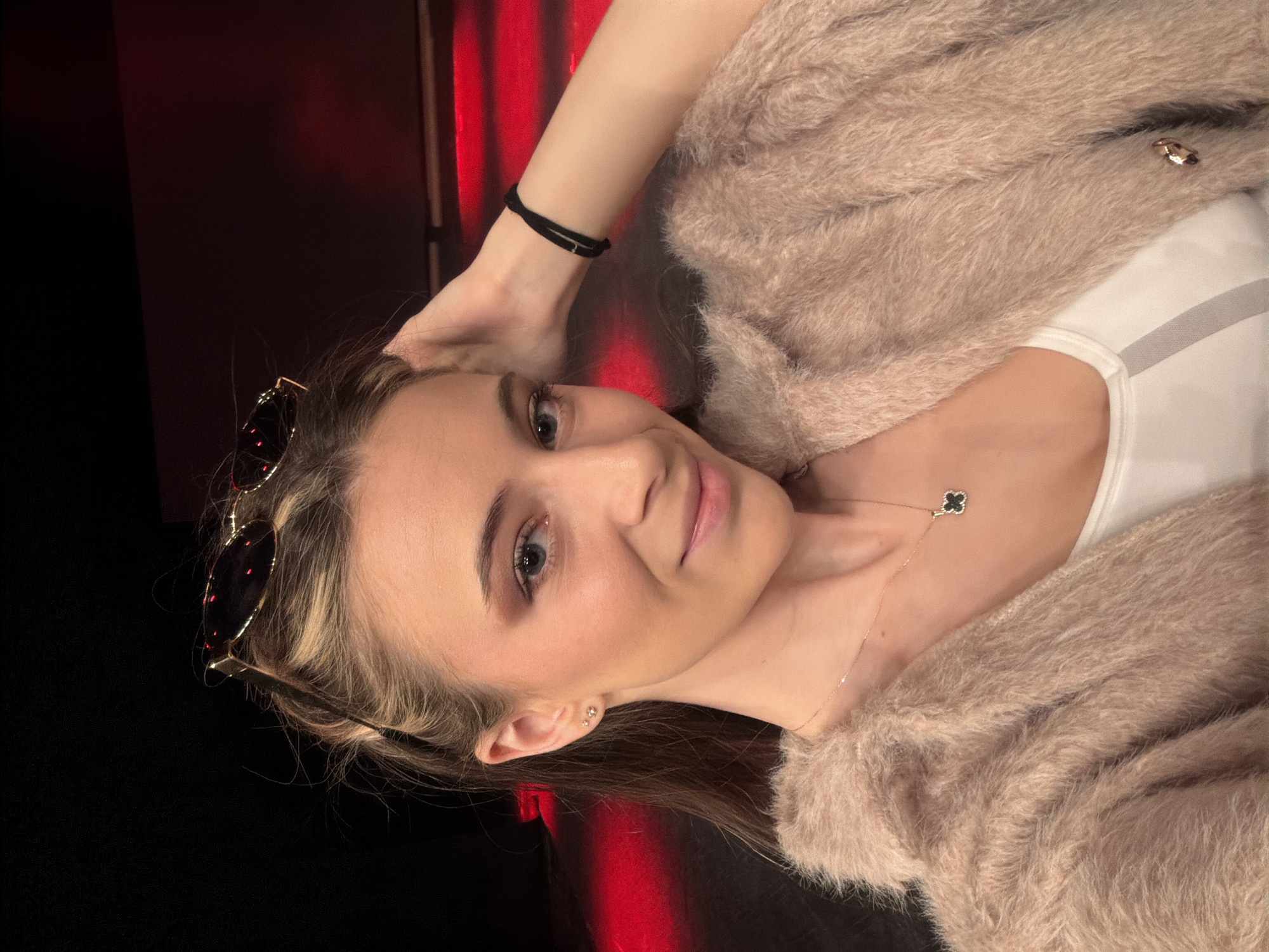 A young woman poses for a selfie. She is fair-skinned with light brown hair, wearing a fluffy pink sweater over a white top and a delicate necklace. Sunglasses are perched on top of her head, and she wears a black bracelet on her wrist. The background appears to be dark with red lighting. The exact location cannot be confirmed to be Plovdiv, Bulgaria from the given image.