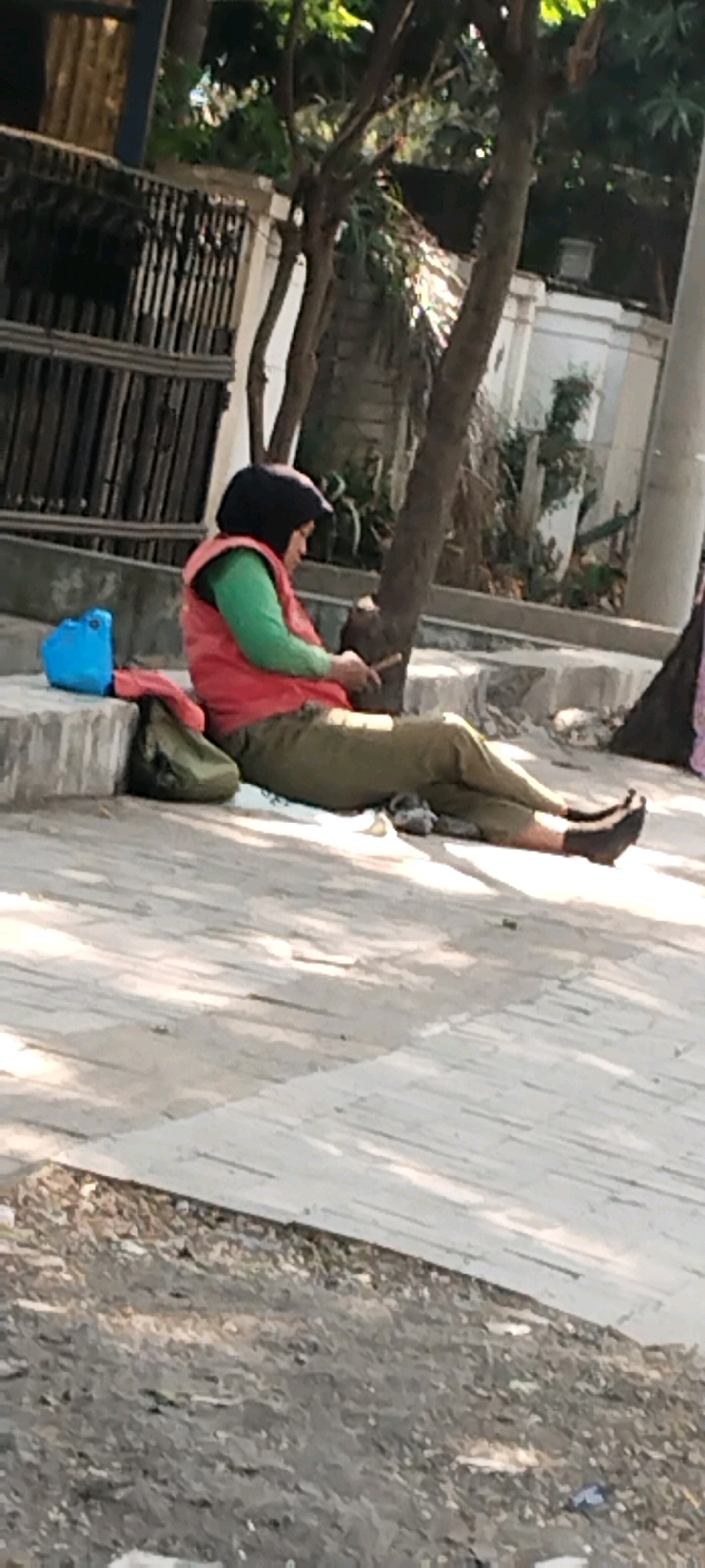 The image depicts a person wearing a dark head covering, a green long-sleeved shirt, a reddish vest, and olive-colored pants. The individual is seated on a concrete ledge, resting their head on their hand and holding a thin, stick-like object. To the left of the person is a bright blue bag and a green backpack. The person is also sitting on a white cloth. The background shows a fence made of vertical wooden planks and trees with dappled sunlight filtering through their leaves. The ground in the foreground is paved with light-colored tiles, with some dirt and gravel visible. The scene appears to be an outdoor public space, possibly a sidewalk or park area in Semarang, Indonesia, given the typical urban environment and vegetation.