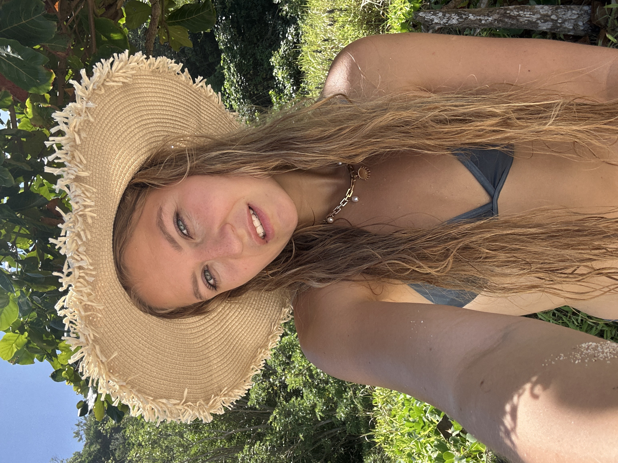 A young woman poses for a selfie outdoors. She is wearing a straw hat with a fringed edge, a dark-colored bikini top, and a gold necklace. Her long, light brown hair is wet. The background consists of green foliage. The location is likely in a tropical setting, possibly Kuta Selatan, Indonesia, given the prompt. There is sand on her arm and shoulder.