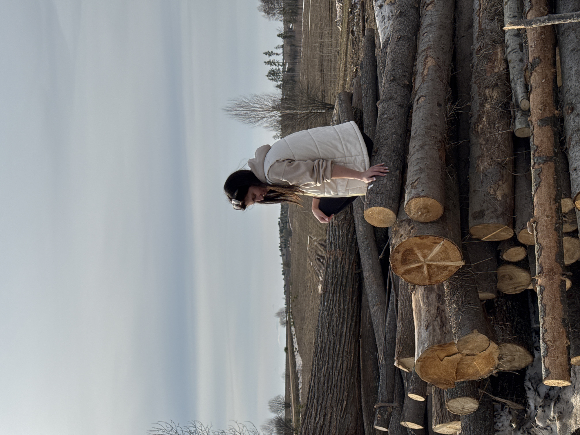 The image shows a stack of logs in an open field, likely a logging site or storage area. A woman is positioned on top of the logs. She has dark hair and is wearing sunglasses, a light-colored hooded jacket, and dark pants. She is bent over, looking down towards the logs. The logs are of varying sizes and appear to be freshly cut. The ground is covered in dry grass and some snow. In the background, there is a field with some trees and a clear sky with some cloud cover. The photo was taken during daylight hours. The location is Van'ki, Russia.