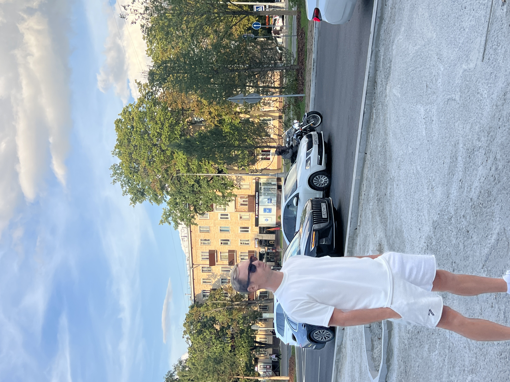 A man is standing on a sidewalk in Moscow, Russia. He is wearing a white t-shirt and white shorts, and sunglasses. In the background, there is a street with cars and buildings. One car is black with a license plate that reads "0822XB 797". A motorcycle is also visible. The sky is blue with some clouds.
