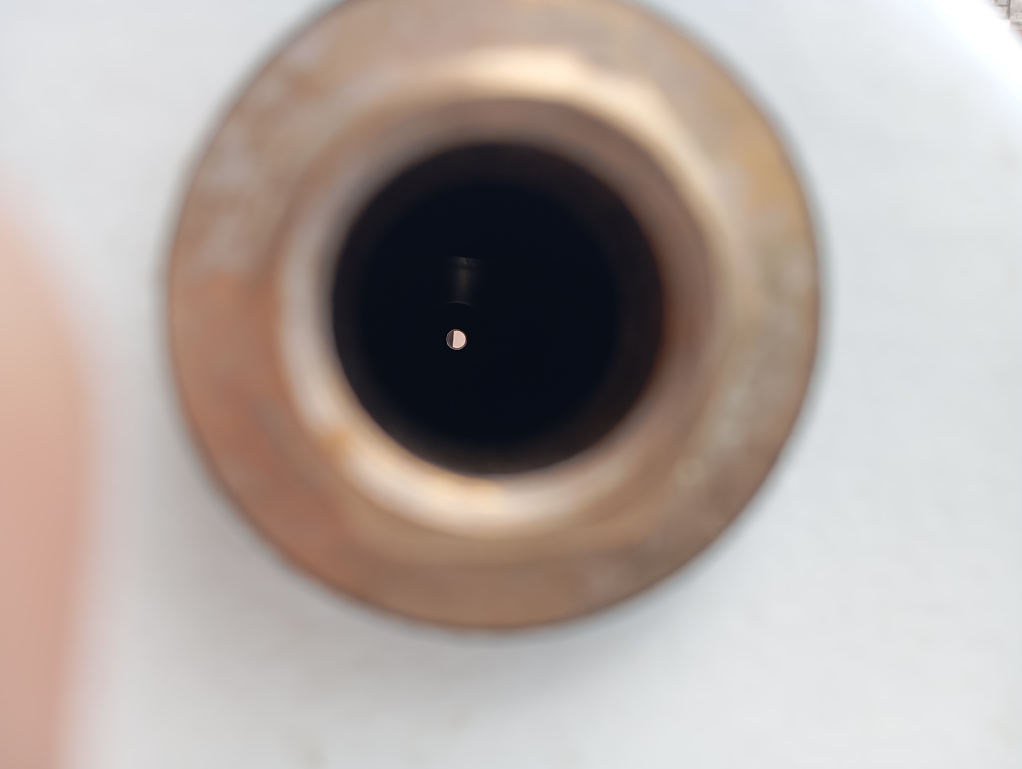 The image is a close-up, blurred view looking into a metal tube. The tube has a copper-colored rim and a dark interior. In the center of the dark interior, a small, circular highlight is visible. The background is out of focus and appears to be a light-colored surface. The scene is not set in a specific location in a city; it is a detail shot of an object.
