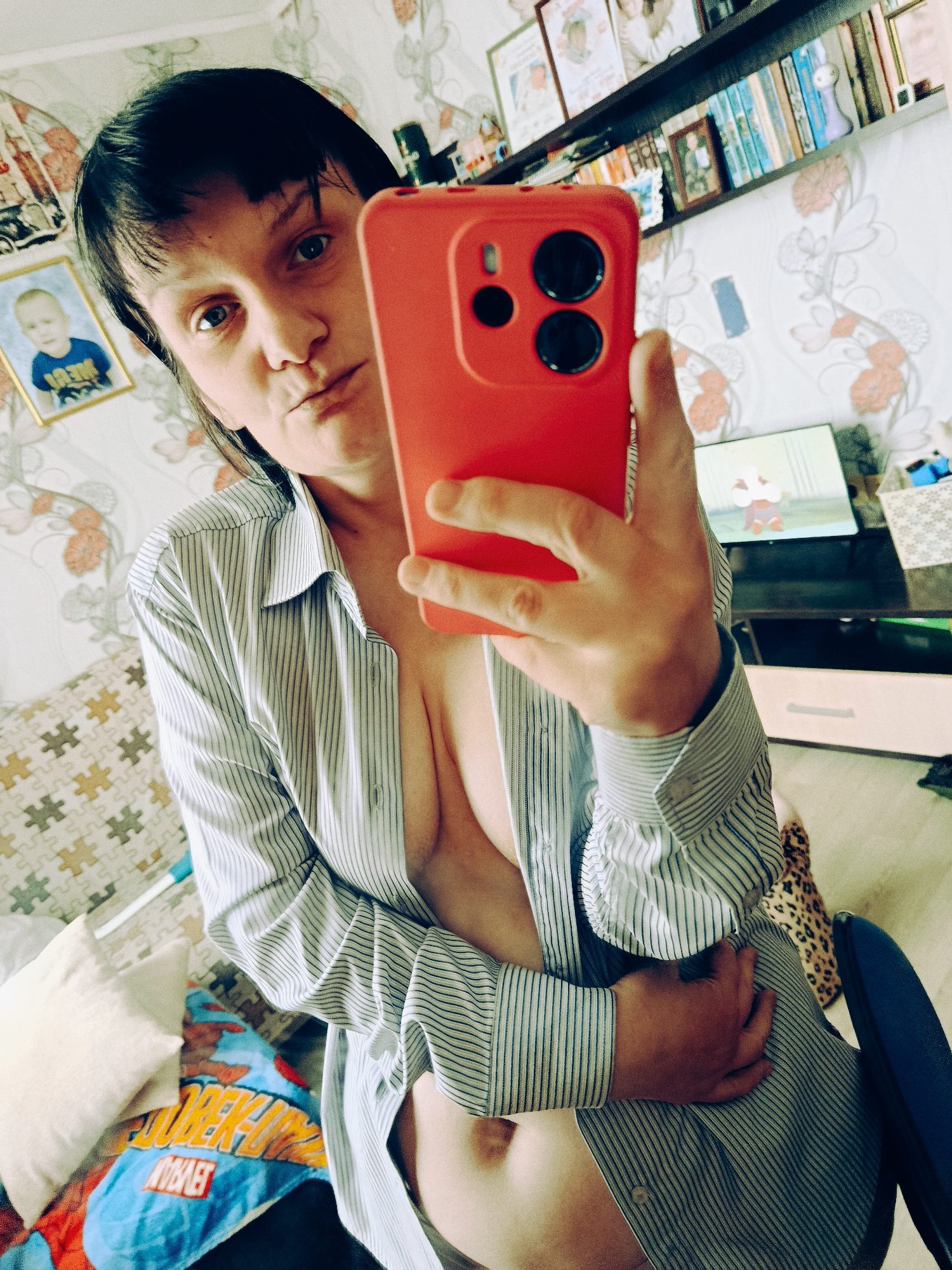 A fair-skinned person with short, dark hair is taking a selfie in what appears to be a home interior. The person is wearing an unbuttoned striped shirt, revealing part of their torso. They are holding a red smartphone with a camera prominently visible. The background includes a wall with floral wallpaper, a shelf filled with books and decorative items, a television screen displaying a cartoon, and various household objects like pillows, a blanket, and a chair. A framed photograph of a child hangs on the wall. There is no verifiable information to confirm this image was taken in Фоки, Russia.
