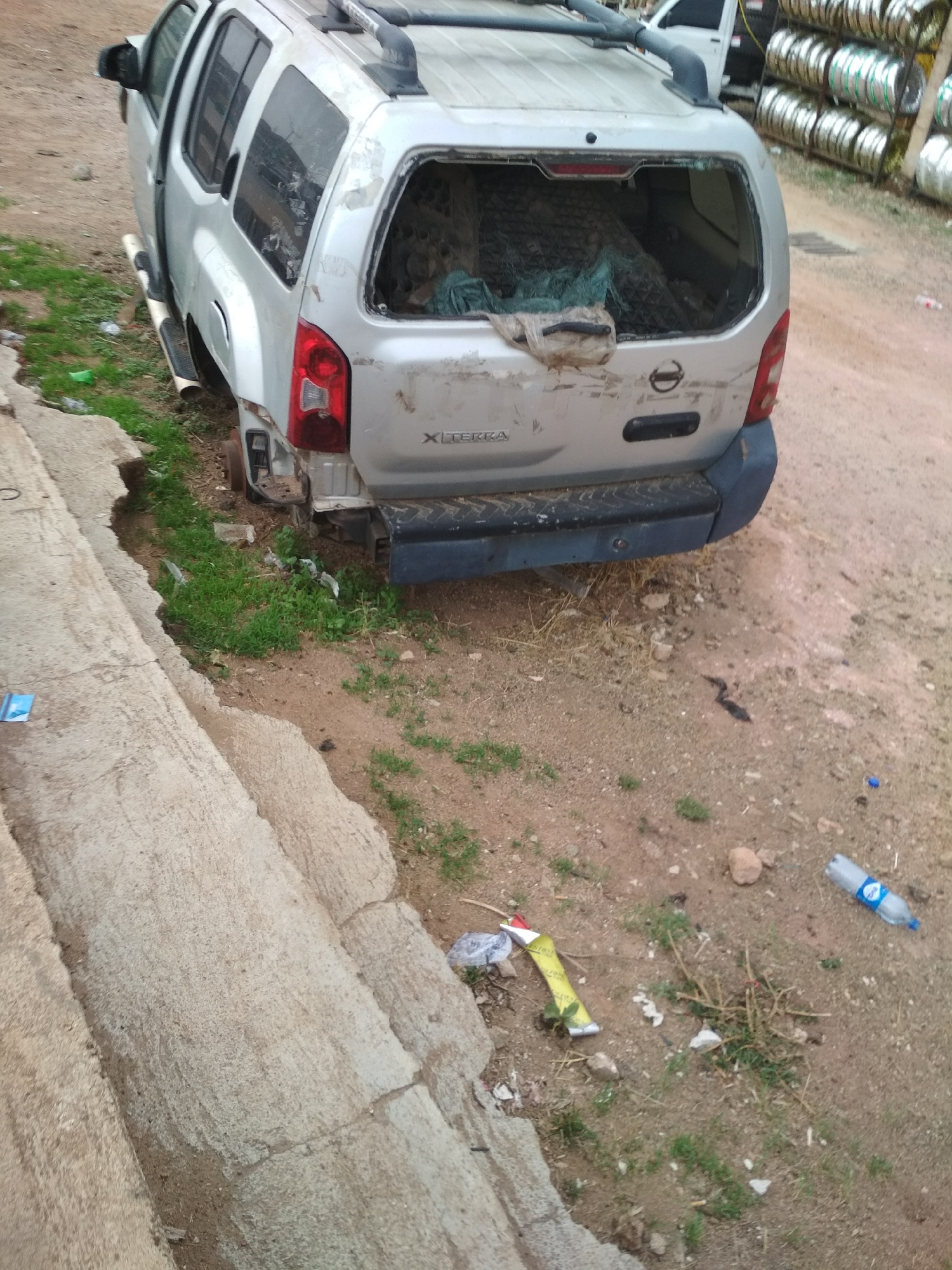The image shows a damaged silver Nissan Xterra SUV with its rear window broken out. The vehicle is missing its rear bumper and is resting on a dirt area with some grass, near a concrete curb or steps. There is debris on the ground near the vehicle, including a water bottle, wrappers, and twigs. In the background to the right, there is a rack of metallic objects, possibly rims. In the distant background to the upper right, there is another vehicle. The scene is located in Gura Top, Nigeria. There are no visible people in the image.