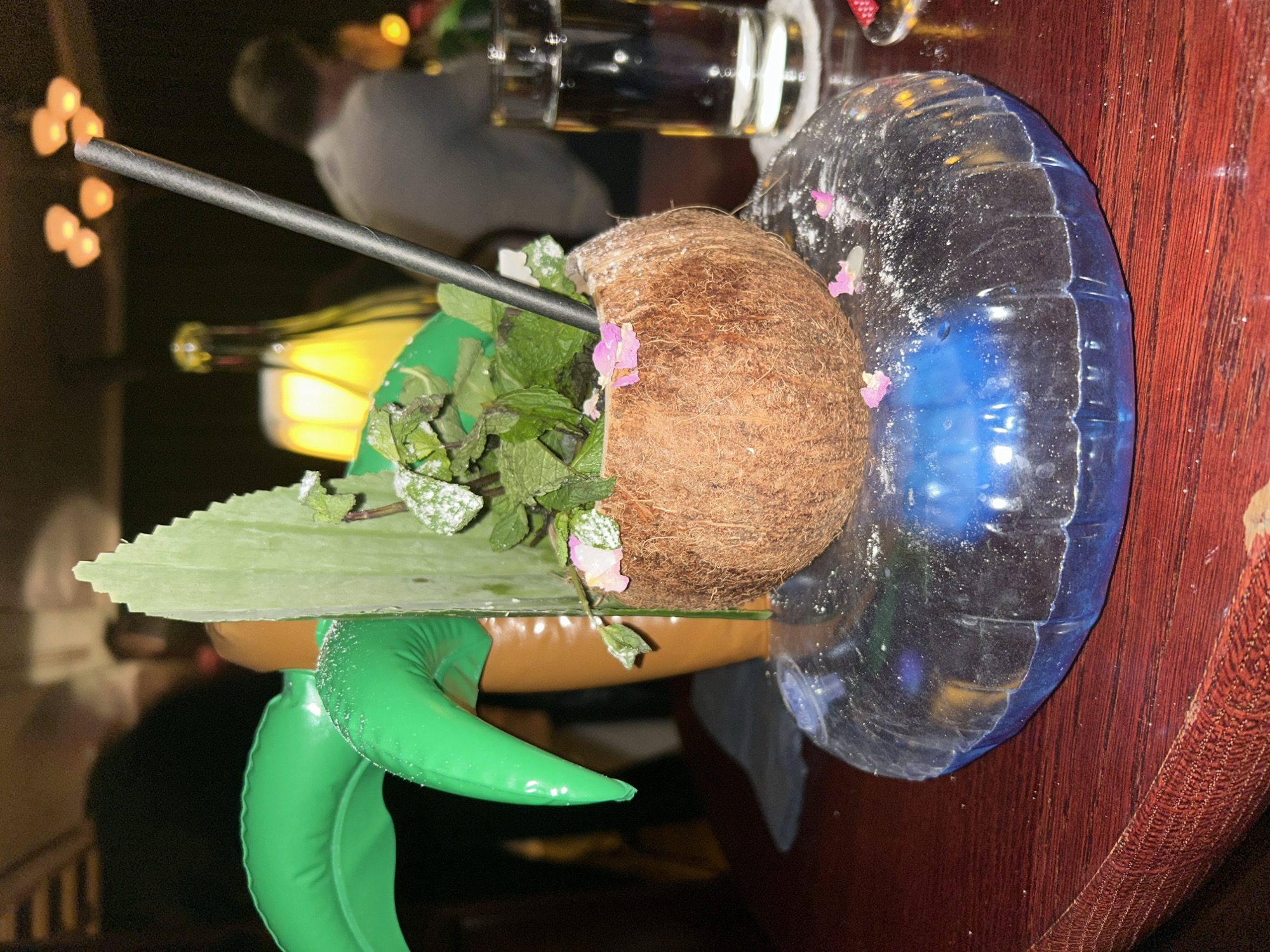 A coconut cocktail is served on a wooden table, perched inside a turtle-shaped inflatable pool toy, in a dimly lit restaurant. The cocktail is topped with fresh mint leaves, decorated with pink flower petals and powdered sugar. The drink is in a halved coconut shell, and has a straw inserted. The inflatable turtle's shell is a light green, and its body is tan. It is sitting in a clear and blue mini-pool inflatable. The table it rests on is a dark, stained wood with an intricate inlay. A glass of a dark beverage is visible in the background, along with blurred human figures. While I cannot ascertain the location of this image, it does not seem possible to determine its exact location to Berlin, Germany, or anywhere else for that matter.