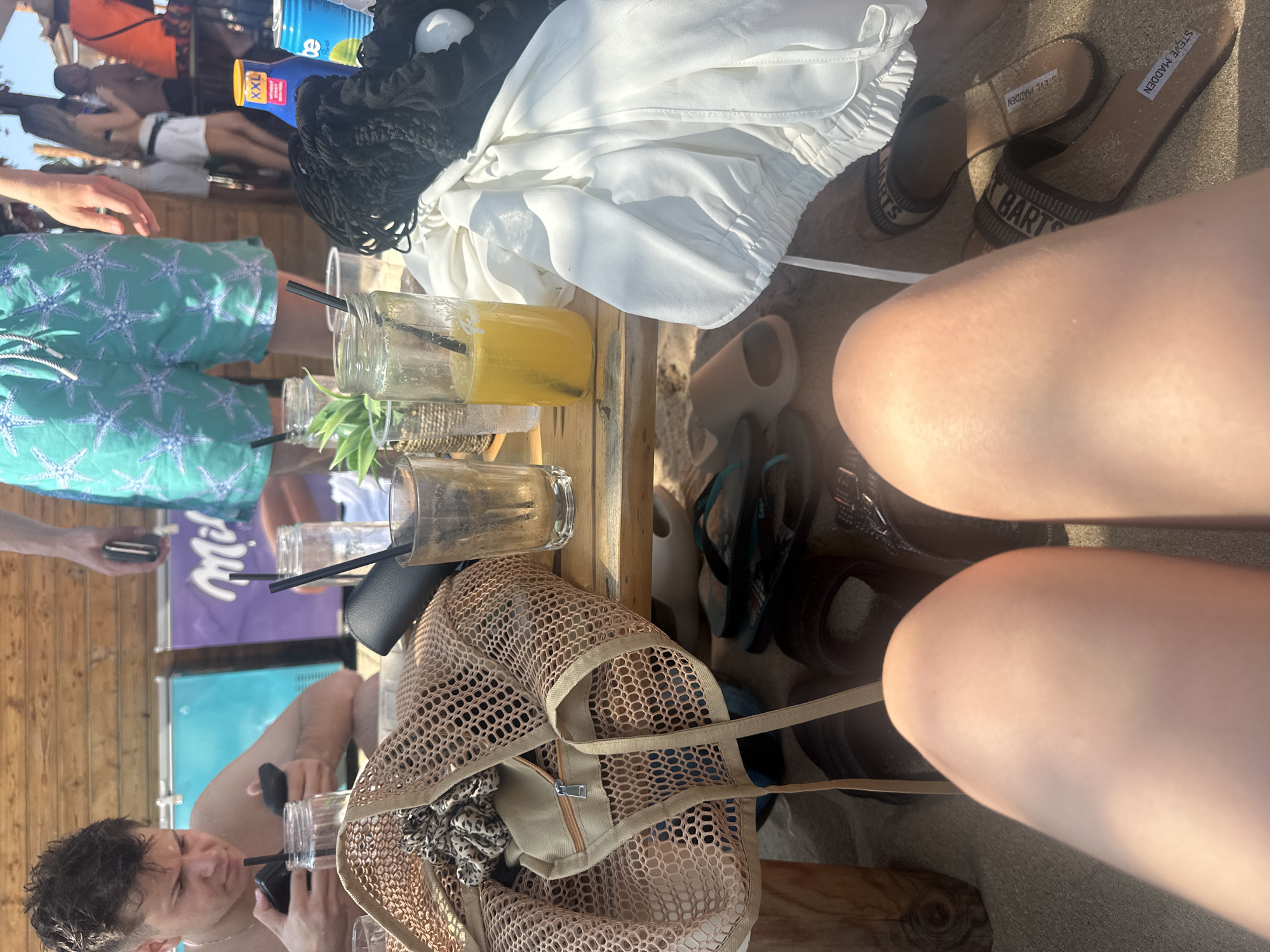 The image depicts a casual gathering, likely at a beach bar or outdoor cafe, given the sandy ground and casual attire. The scene is set during the day, with bright sunlight casting shadows, suggesting fair weather. Several people are present, though their faces are not entirely clear or are partially obscured. One person, a young man, is visible in the lower portion of the image, holding a drink with a straw and looking downwards. He is wearing a necklace. Nearby, a mesh bag rests on a wooden surface, revealing some patterned fabric inside.
On the wooden table, several drinks in glass jars with straws are visible. One drink is a vibrant yellow. Also on the table are a white jacket or cover-up and a black drawstring bag. Further into the background, another person is seen wearing blue and green patterned shorts. A pair of tan slide sandals with the brand name "STEVE MADDEN" clearly visible are on the sand. The overall atmosphere appears relaxed and social, consistent with a vacation or leisure activity.
Text visible in the image includes "STEVE MADDEN" on the sandals. There are also faint labels on what appear to be drink bottles or cans in the background, but they are largely illegible. The general setting is consistent with a coastal location like Sozopol, Bulgaria, which is known for its beaches and summer tourism.