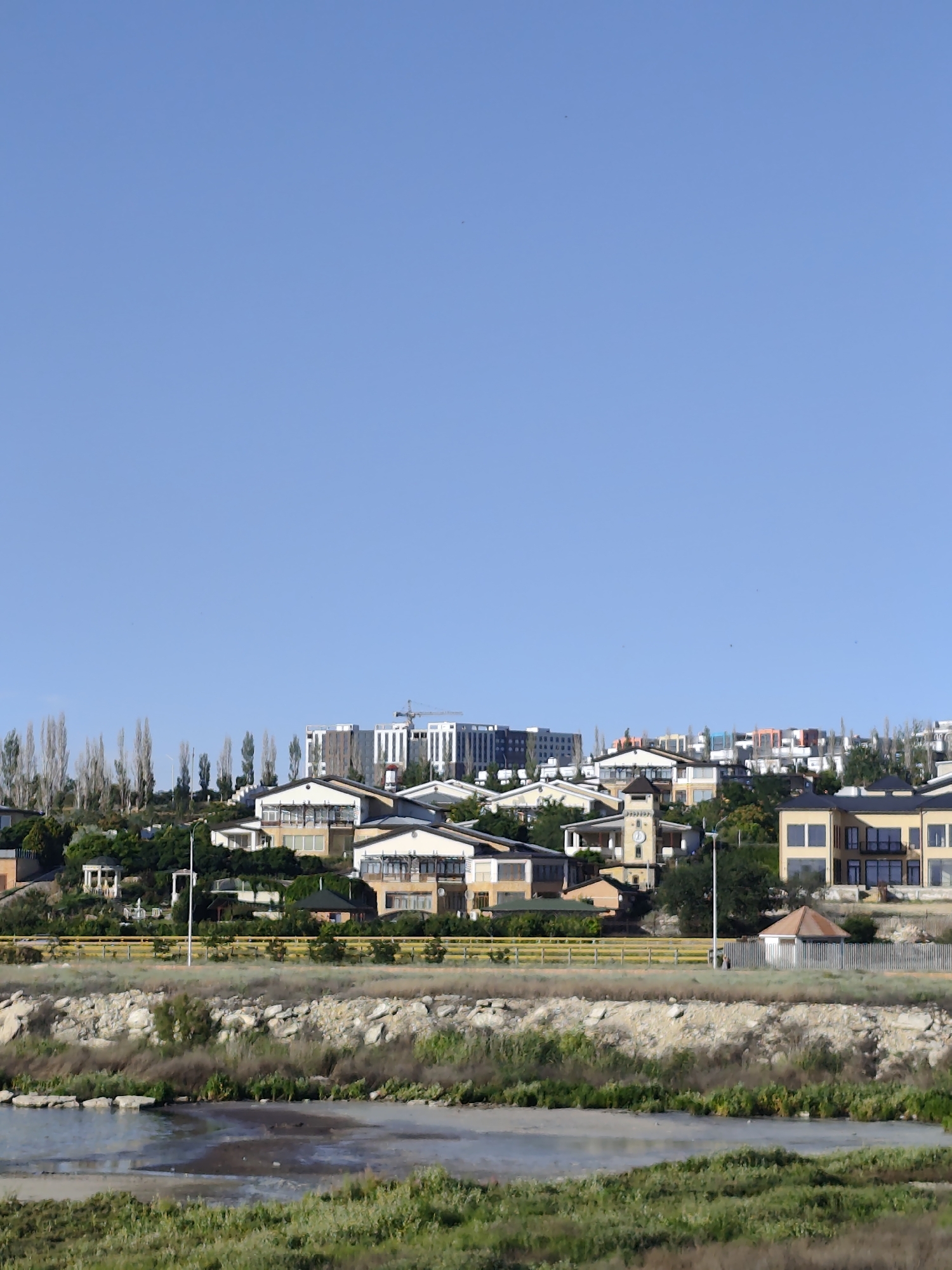 The image depicts a coastal hillside residential area under a clear blue sky, suggesting fair weather and daytime. The scene is dominated by a collection of modern houses with varied architectural styles, many featuring large windows and balconies. Some houses are built in a cluster, while others are more spread out. In the background, a large, multi-story building with a construction crane suggests ongoing development.
In the middle ground, a yellow fence runs horizontally across the image, separating the residential area from the foreground. Below the fence, a strip of rocky terrain is visible, followed by a body of water, possibly a lagoon or a shallow bay, with patches of green vegetation growing along its edges. The water appears calm, with some muddy areas. The foreground consists of more lush green grass and vegetation.
Notable details include a tall lamppost near the center of the image, illuminating the area and suggesting that this is a residential street or public space. Several tall, slender trees are planted in a row on the hillside, adding to the landscape. The overall impression is one of a developing suburban or coastal community. No people are visible in the image, and no text is present. The location context of Aktau, Kazakhstan, implies a possible arid or semi-arid climate, which might explain the dry appearance of some vegetation and the rocky terrain.