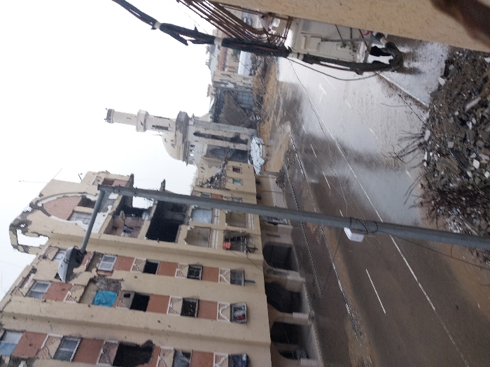 The image depicts a heavily damaged urban street scene in Khan Yunis, Palestinian Territory. In the foreground, a gray streetlamp stands prominently on the left. To its left, a multi-story building, likely residential, exhibits extensive damage, including large sections of its facade missing, numerous shattered or absent windows revealing dark interiors, and visible structural compromise. The building's exterior is beige with brown horizontal banding and decorative arches. A blue tarp hangs from one upper-story window opening.
The asphalt road running through the center of the image appears wet, possibly from rain, reflecting the overcast sky. White lane markings are visible on the road surface. Extensive rubble and debris, consisting of shattered concrete, metal fragments, and exposed rebar, line the street and are particularly concentrated in the immediate foreground, obscuring the ground level.
In the mid-ground, a mosque with a tall minaret and a dome shows significant structural damage. Part of the mosque's dome appears collapsed, and sections of its walls are damaged or missing. Further down the street, beyond the mosque, more damaged buildings and widespread debris are visible. On the right side of the image, the facade of another building is visible, with dark electrical conduits running vertically down its surface. No individuals are visible in the scene. The overall environment conveys widespread destruction.