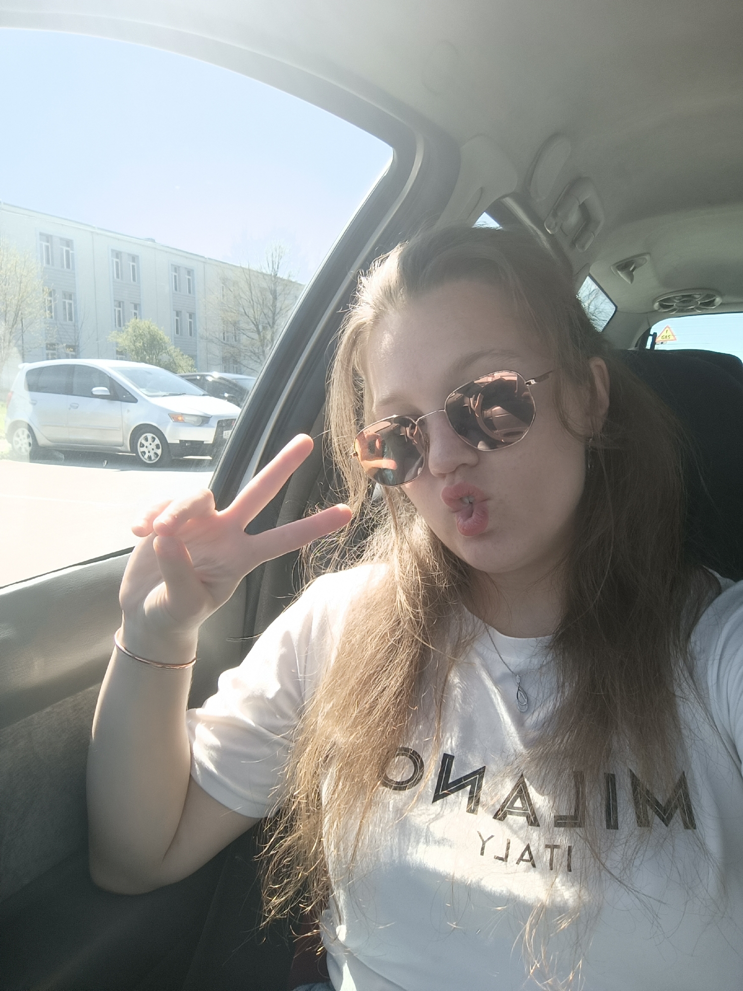 A young woman is in the front seat of a car, making a peace sign with her left hand and pursing her lips. She is wearing rose-tinted sunglasses and a white t-shirt with "MILANO ITALY" printed on it. Her hair is long and light brown. Outside the car window, a silver car is parked in front of a multi-story building. Based on the location of the "GAS" triangle visible on the upper right corner, the scene likely takes place in Plovdiv, Bulgaria, where such markers are prevalent near gas stations.