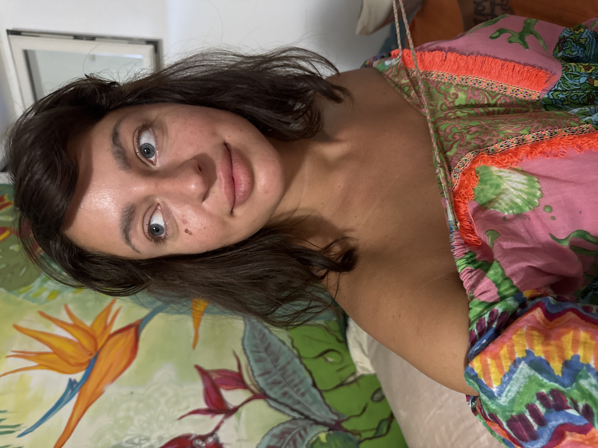 A young woman with dark, wavy hair is looking up at the camera, her head tilted to the side. She has wide, blue eyes, a small nose, and full lips. A few freckles are visible on her cheek. She is wearing a brightly colored, patterned dress with an off-the-shoulder neckline and fringed trim. The dress features a mix of pink, green, orange, yellow, and blue hues, with floral and abstract designs.
The background is blurred but appears to be an indoor setting, possibly a bedroom or living room. There is a large, colorful piece of artwork or fabric behind her, depicting large tropical flowers, including a bird of paradise, in vibrant shades of orange, yellow, and green. A white framed mirror is visible in the upper left corner of the image.
The lighting suggests it could be daytime, possibly late afternoon or evening, given the soft, diffused light. The woman's expression is neutral or slightly curious. The image is a selfie, taken from a close-up perspective. The overall impression is casual and relaxed, possibly taken at home in San Benedetto del Tronto, Italy.