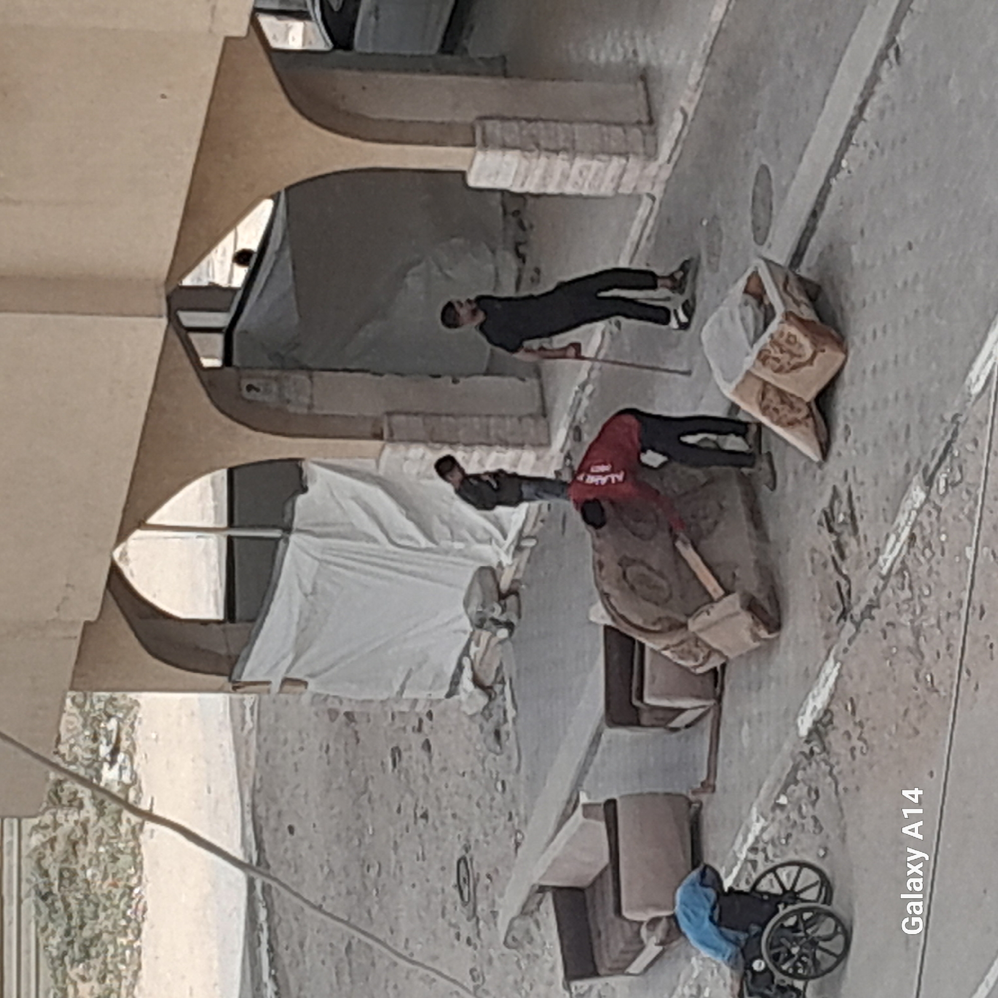 A ground-level, medium shot depicts a street scene of Unknown, Unknown. There are multiple individuals present, including one person wearing black clothing and another in a red shirt. They appear to be situated in front of an arched building or structure. Various objects such as furniture pieces (including what seems to be a couch and possibly other upholstered items) are scattered across the scene. A wheelchair is visible near the bottom right corner of the shot. The overall setting gives the impression of an urban area, potentially depicting the aftermath of displacement or movement of belongings.