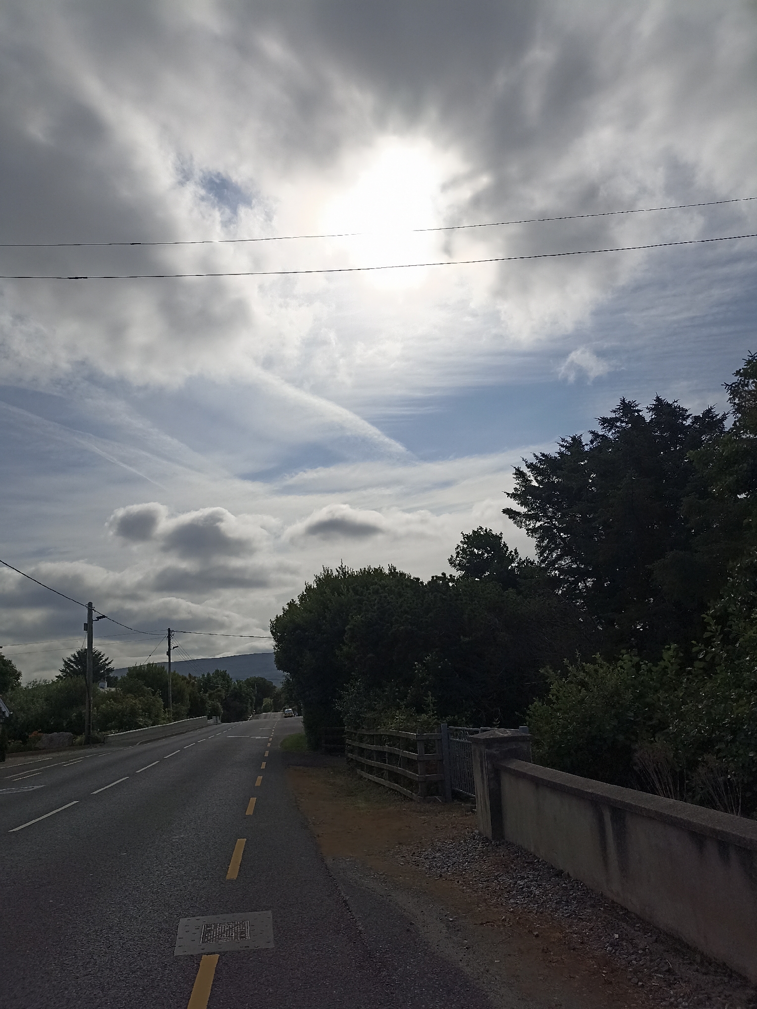 The image shows a road in Annagry, Ireland, on a partly cloudy day. The road has a yellow dashed line down the center and a solid white line on the left side. There is a manhole cover in the road with a yellow line going over it. On the right side of the road, there is a stone wall and a wooden fence. Trees and bushes line the side of the road. In the distance, there are hills. The sky is filled with clouds, and the sun is partially visible. There are power lines running across the sky.