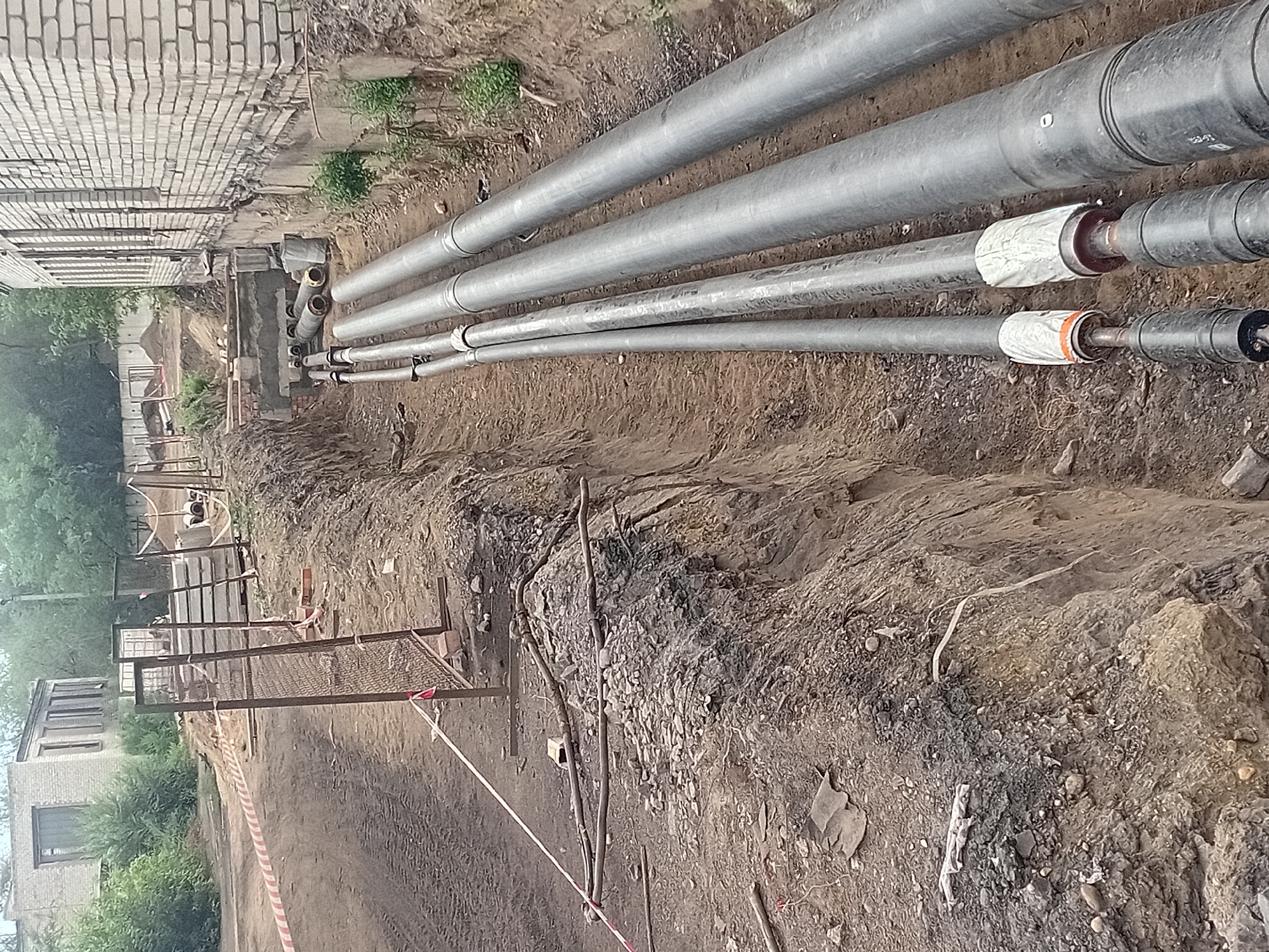 The image depicts a construction site in the city of Semey, Kazakhstan, focusing on an open trench where large diameter pipes are being laid. The trench runs alongside a brick building and a dirt road. Several grey pipes, some with protective coverings at their joints, are visible within the trench. The surrounding environment shows signs of excavation, with piles of dirt and exposed earth. A red and white striped caution tape is strung across the scene, indicating a work area. The overall impression is one of ongoing infrastructure development.