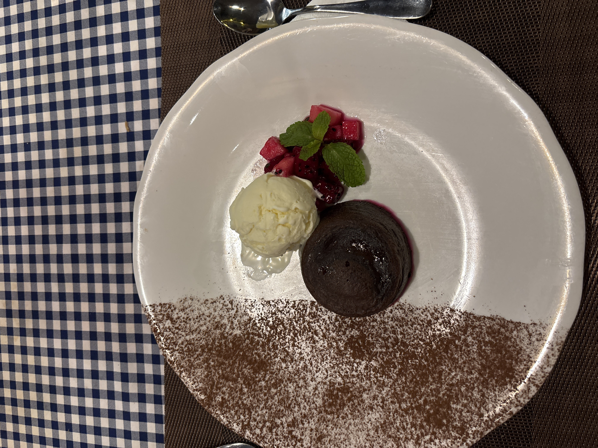 A top-down shot shows a dessert on a white plate, consisting of a scoop of vanilla ice cream, a small pile of diced red fruit and mint leaves, and a chocolate lava cake with cocoa powder sprinkled on the plate below. The plate is positioned on a dark brown surface, partially overlaid by a blue and white checkered tablecloth. A silver spoon is placed towards the upper part of the plate. The image appears to be taken in a restaurant setting. It is unknown whether it is located in Đà Nẵng, Vietnam.