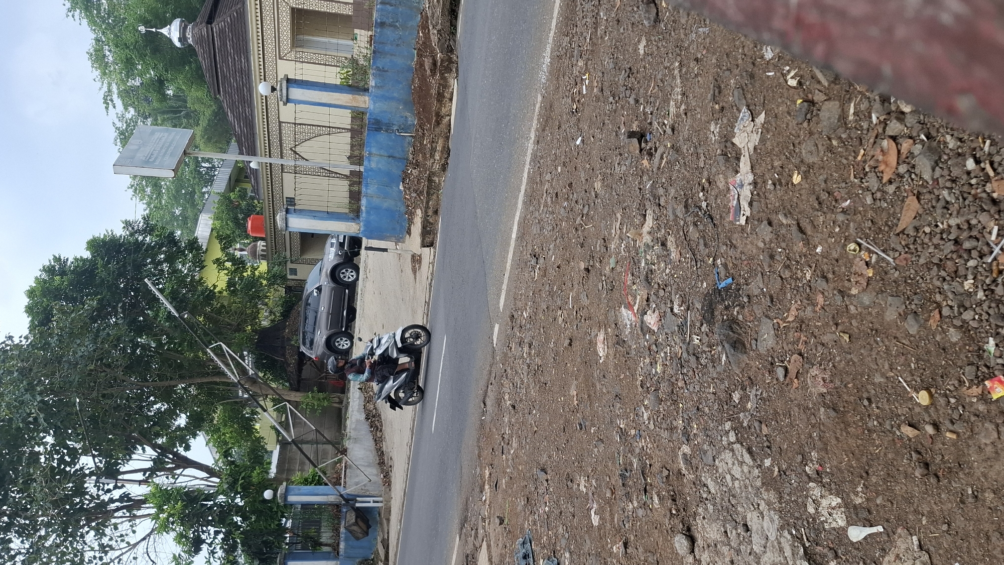 In Cilaku, Indonesia, a motorcyclist wearing a helmet rides a silver scooter on a paved road. The road is bordered by a blue and white fence. Behind the fence, a dark gray SUV is parked. A building with a patterned facade and a dome is visible. To the left of the building, a yellow structure stands. A tall, metallic pole leans precariously over the scene. The ground in the foreground is rough and covered with dirt, rocks, and scattered debris. The sky is overcast.