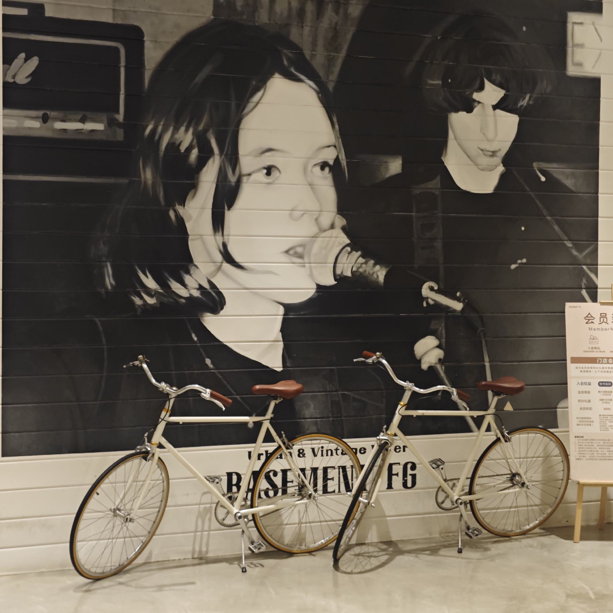 The image shows two vintage bicycles parked in front of a black and white mural depicting two people singing into microphones. The mural appears to be painted on a wall made of horizontal planks. In the background, a Marshall amplifier is visible. The floor is a light grey color. A sign is also visible to the right, in Chinese, with the English translation "Member Notice" at the top. The bicycles are light-colored with brown seats and handlebars. The mural and bicycles are located in the Liangjiang New Area of Chongqing, China. There is a sign that says "Urban & Vintage basement FG" which may indicate a store name or location.