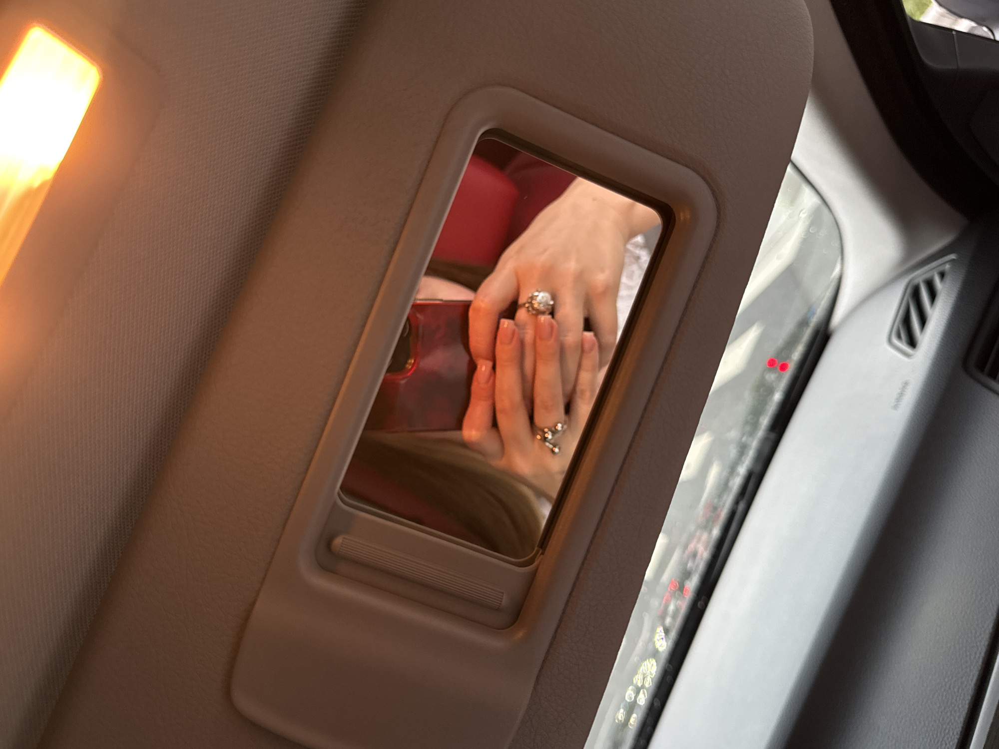 The image is a close-up, high-angle shot of a car's interior, focusing on a vanity mirror in the sun visor. Reflected in the mirror is a person's hands holding a red smartphone, which is capturing the reflection. The person is wearing a prominent ring on their left hand and appears to have manicured nails. The reflection also shows a glimpse of blonde hair and a red garment. Outside the car, visible through the windshield, is a blurry street scene with traffic lights, indicating the car is likely in motion or stopped in traffic. The interior of the car is dimly lit, with the exception of a bright overhead light that casts an orange glow. The setting is not identifiable as Yiwu, China, or any specific city.
