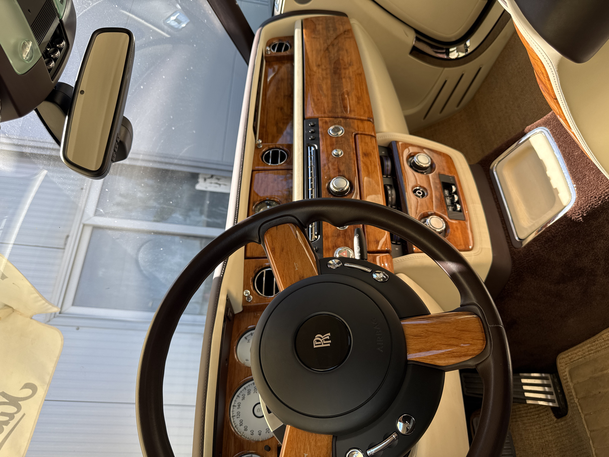 The image shows the interior of a luxury car, likely a Rolls-Royce, with a focus on the steering wheel and dashboard. The steering wheel is dark brown leather with wood accents and the Rolls-Royce emblem in the center. The dashboard is adorned with polished wood veneer and features various buttons and controls. The interior appears to be in pristine condition, suggesting careful maintenance or a new vehicle. The surrounding environment is a light-colored, possibly metallic, textured wall with a window or door in the background, hinting at an indoor setting such as a garage or showroom. The specific location of Dub, Montenegro, cannot be determined from the visual information provided in the image.