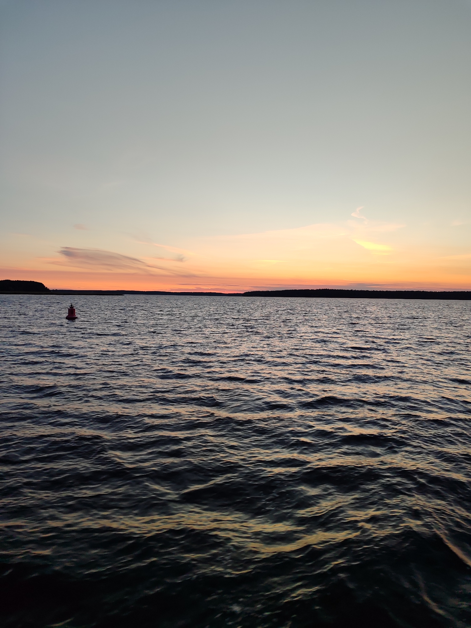 A serene sunset over a body of water features a calm, rippling surface reflecting the warm hues of the sky. A solitary red buoy bobs gently in the foreground, marking the vast expanse of the water. The horizon is lined with a dark, silhouetted forest, creating a stark contrast against the gradient of the twilight sky. The sky transitions from a pale blue at the top to soft oranges and pinks near the horizon, with wispy clouds adding texture to the celestial canvas. The scene is peaceful and undisturbed, conveying a sense of quiet solitude.