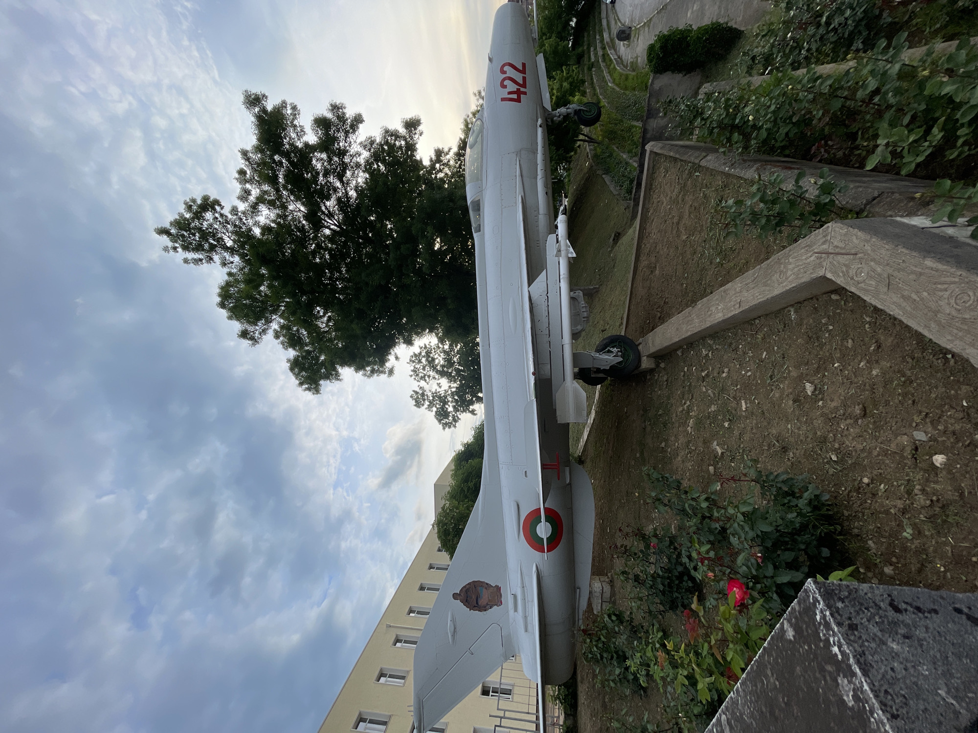 The image shows a MiG-21 fighter jet on display in a garden in Unknown, Unknown. The aircraft is painted gray and has the number "422" in red on its fuselage. It is mounted on a concrete plinth in the garden, which also features trees, bushes, and rose bushes. A building is visible in the background. The sky is cloudy.