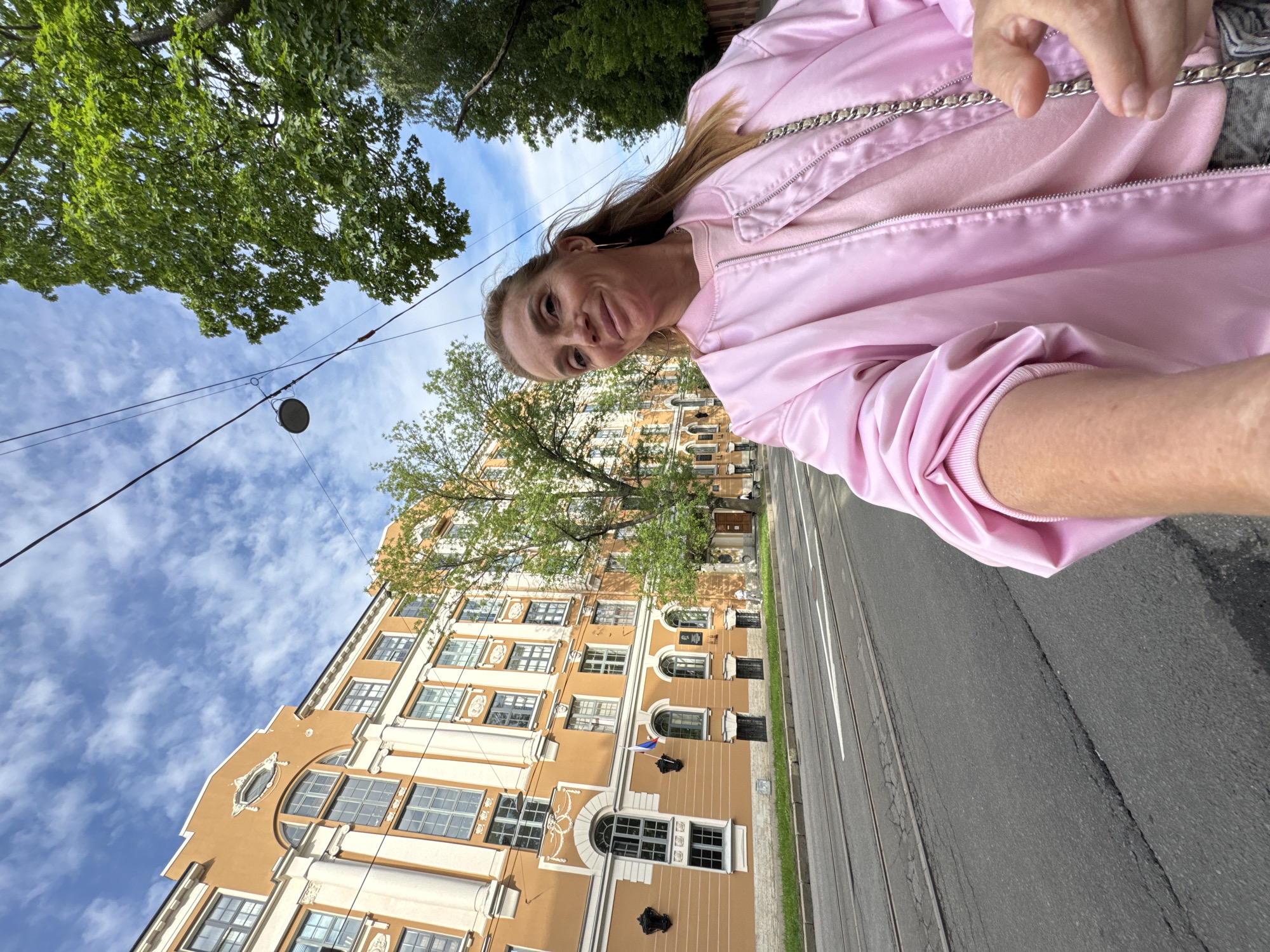 The image depicts a woman taking a selfie outdoors in Saint Petersburg, Russia. She is wearing a pink bomber jacket over a pink sweater and is smiling at the camera. Behind her, a large, orange, multi-story building with numerous windows and architectural detailing lines a street. Tram tracks are visible in the foreground, indicating a public thoroughfare. Lush green trees provide a backdrop and frame the top right of the image, and a partly cloudy blue sky is overhead. The overall impression is a bright, daytime scene in an urban environment.