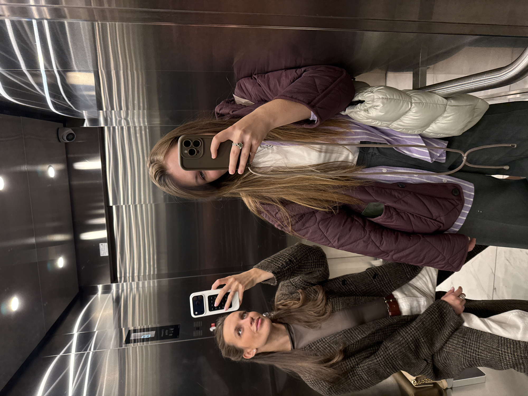 Two women take mirror selfies in an elevator. One woman wears a brown quilted jacket, a purple striped shirt, and dark pants. She holds a cell phone with multiple camera lenses. The other woman wears a brown plaid coat and a brown shirt. She holds a white cell phone. The elevator has a stainless steel interior. Based on metadata, the location is Belgrade, Serbia.
