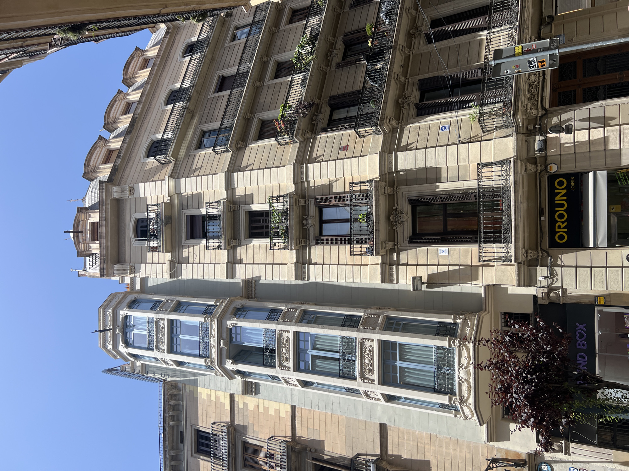 The image shows a street scene in Barcelona, Spain, with a focus on the facades of several tall, ornate buildings under a clear blue sky. The buildings exhibit classic European architectural styles with detailed stonework, balconies with intricate wrought-iron railings, and numerous windows. One particularly prominent building features a striking bay window that extends upwards, with pale blue curtains visible behind the glass. The lower floors of the buildings house various shops, including one with a purple illuminated sign that reads "SECOND BOX," and another with a yellow sign that says "OROUND JOIERS". A small, dark-leafed tree is positioned in front of one of the shops. The scene is captured from a low angle, looking up at the buildings. Sunlight casts shadows on the facades, indicating a bright, sunny day.