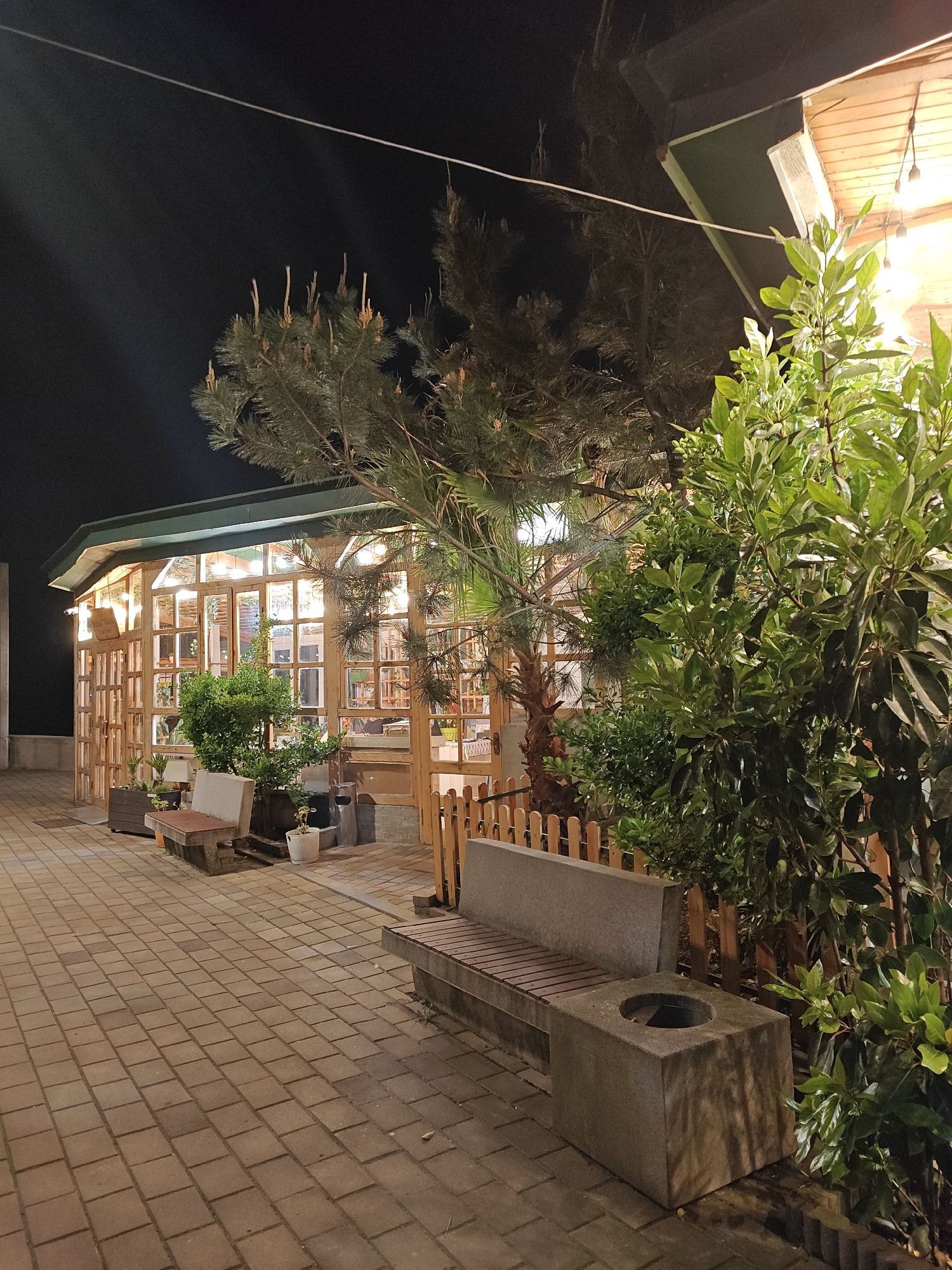Here is a detailed description of the image:
The image shows an outdoor area at night, likely a patio or garden adjacent to a building. The ground is paved with rectangular bricks. The main structure visible is a wooden-framed building with many windows, illuminated from the inside with string lights. In front of this building, there are various plants and trees, casting shadows and adding to the ambiance. There are also benches for seating, one of which has a built-in planter or waste receptacle. A small wooden fence separates the seating area from the vegetation. The sky is dark, indicating it is nighttime. The exact city and location are not identifiable from the image.