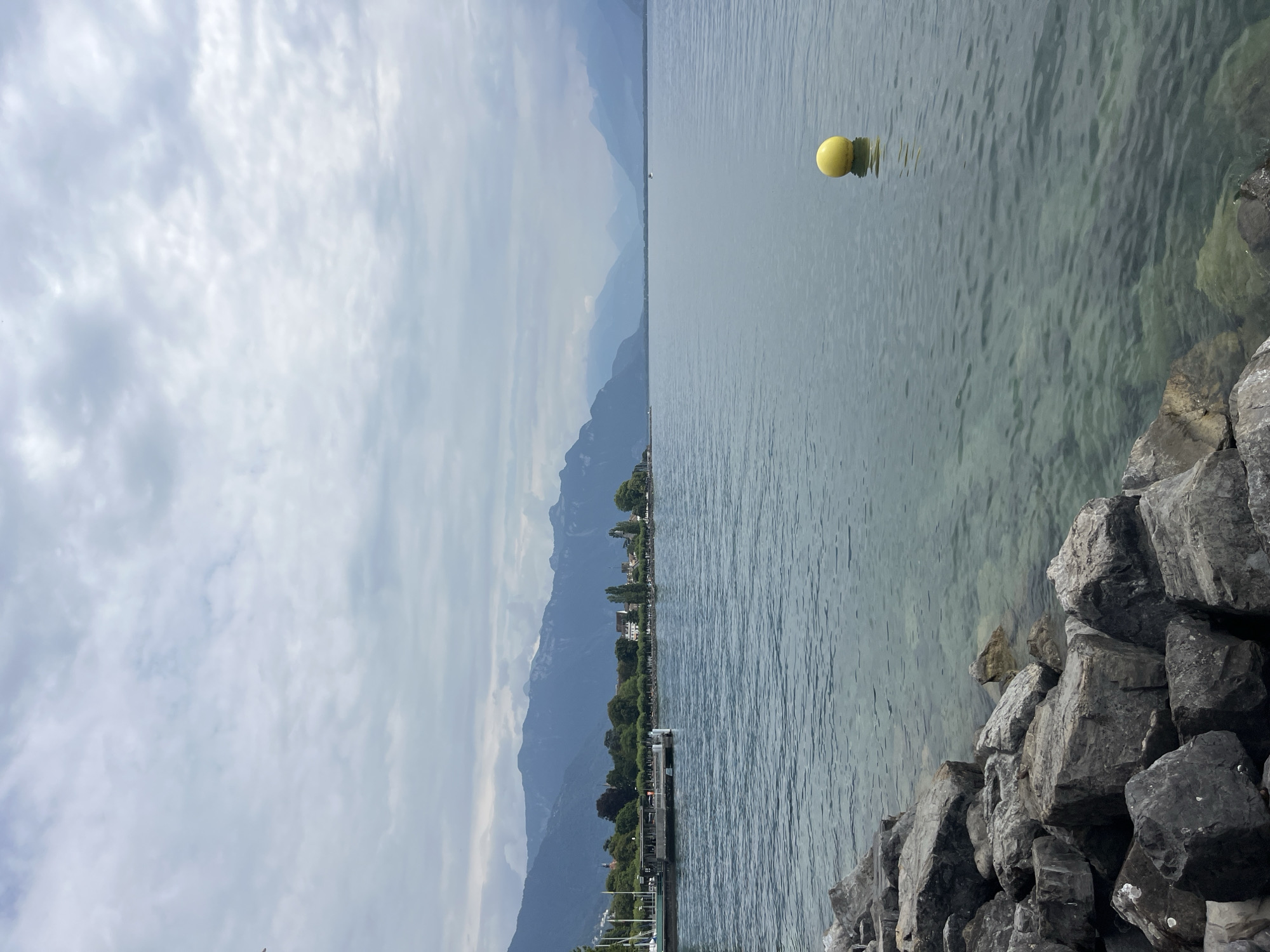 This image depicts a serene waterfront scene in Vevey, Switzerland. In the foreground, a rocky shoreline leads to clear, calm water that transitions to a deeper blue. A bright yellow buoy floats on the water's surface in the mid-ground. Across the water, a line of trees and buildings forms the town's edge, backed by a majestic mountain range under a cloudy sky. The overall impression is one of natural beauty and tranquility.