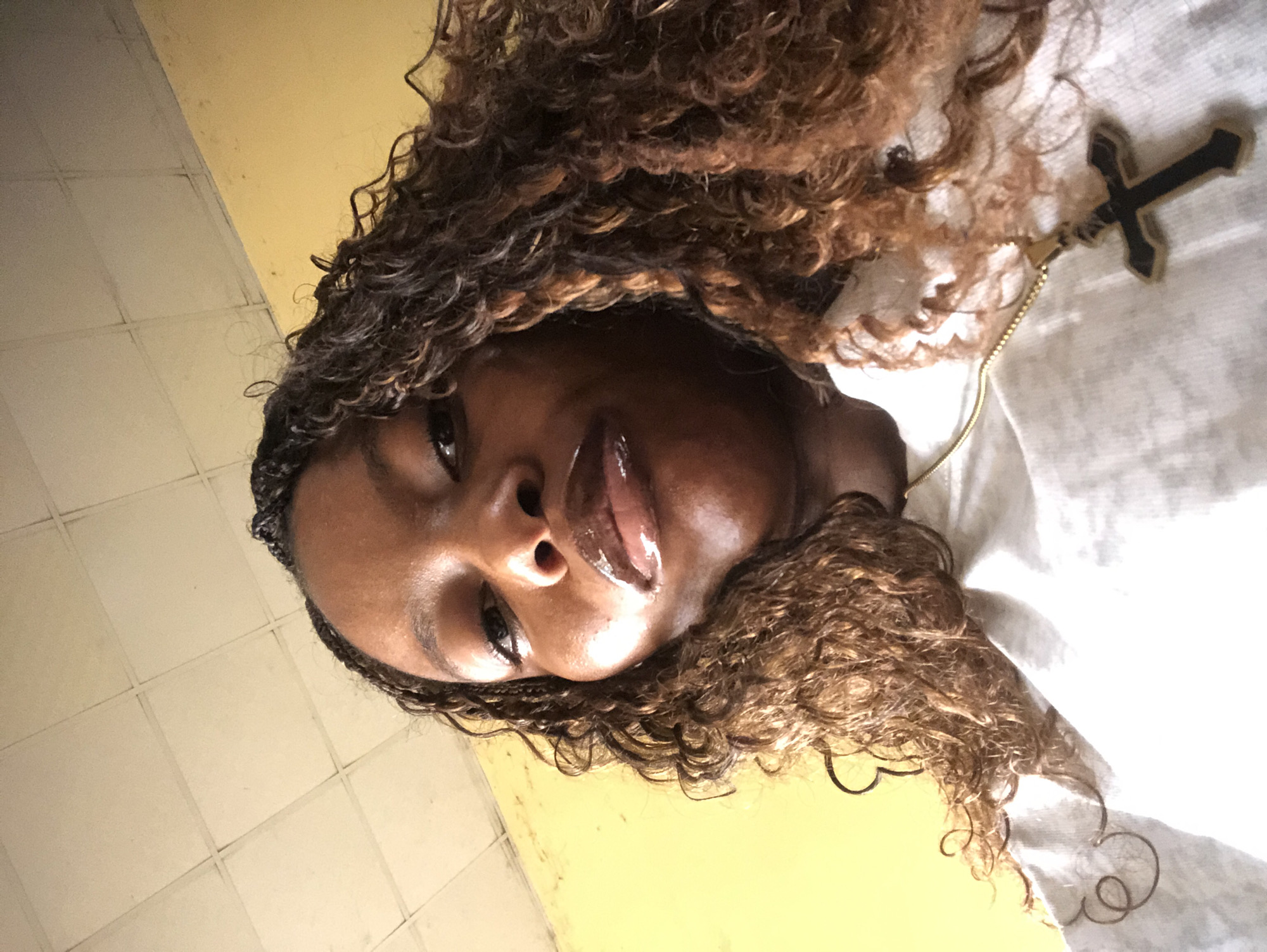 A person with braided hair and curly brown locks is seen in an indoor environment with light yellow walls. A cross necklace is visible. The location is unknown.