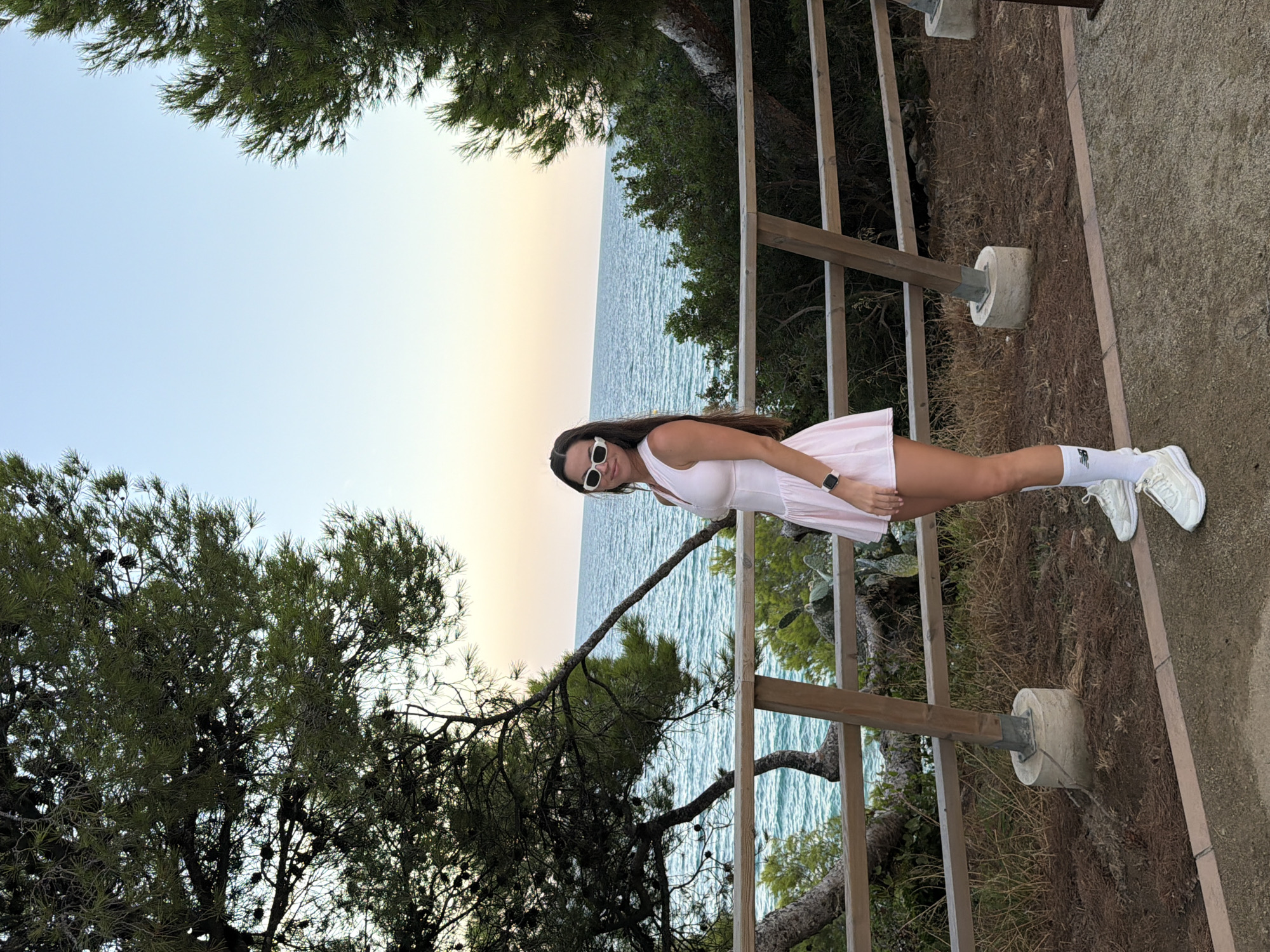 A full-body, eye-level shot shows a woman standing outdoors in Mont-roig del Camp, Spain, at dusk. She is facing the camera, slightly angled to her left, with her head tilted to her right and her gaze directed at the camera. The woman has long, dark hair and is wearing white-rimmed sunglasses, a pale pink sleeveless dress, white athletic socks, and white sneakers. Her left hand rests on her hip, and her right arm is at her side.
The background features a view of the sea, with a wooden railing and fence separating the viewer from the water. Trees with dense green foliage and pinecones frame the scene on either side, with the sky above transitioning from a pale blue to a soft orange as the sun sets. The ground in the foreground is a mixture of gravel and pine needles.