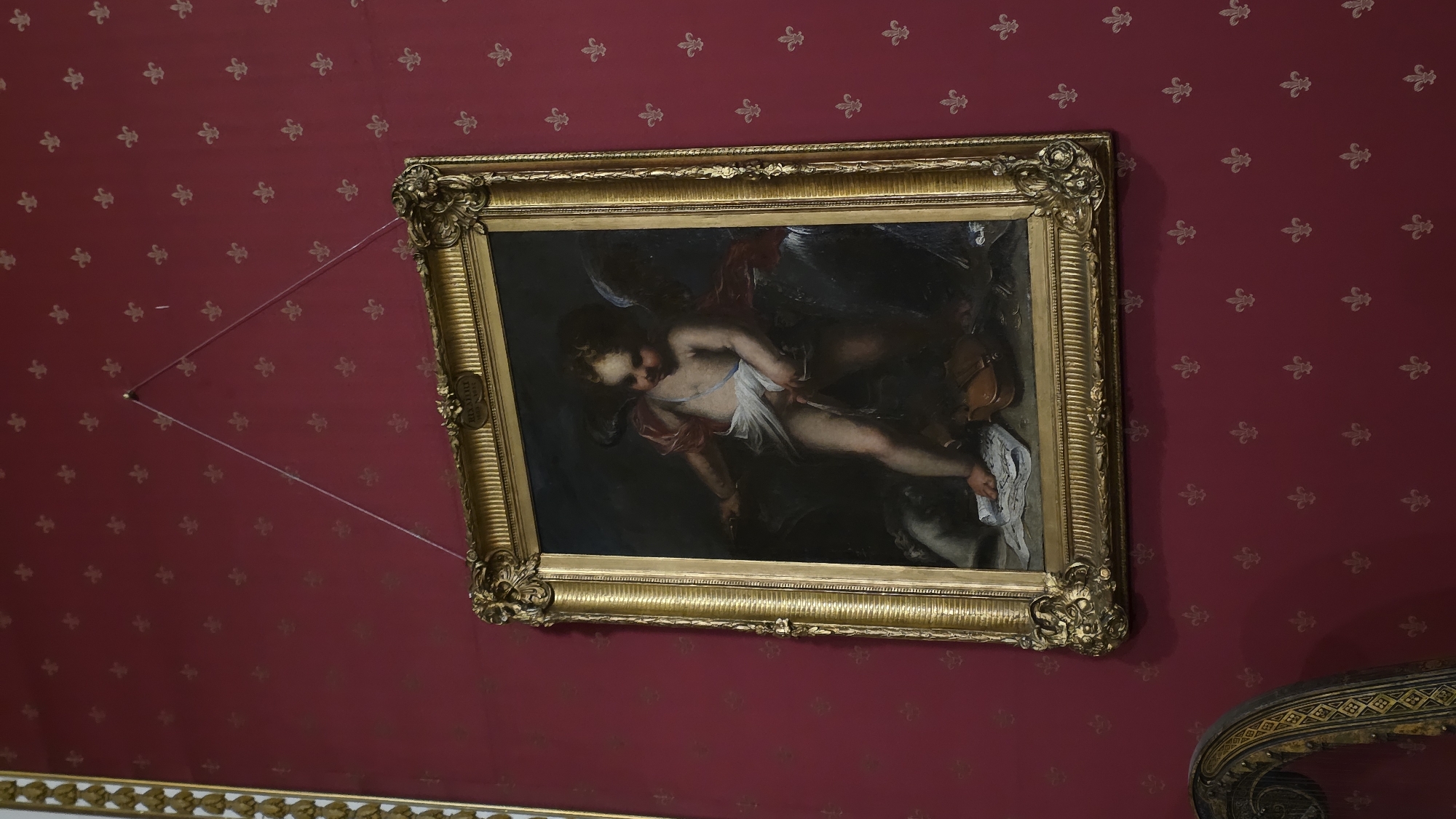 A painting of a cherub, possibly with wings, stands in the center of the frame, depicted in classical style. The cherub is draped in white cloth and appears to be holding something in its right hand. In front of the cherub, a violin and a piece of sheet music are visible on the floor. The painting is housed in an ornate gold frame and hangs on a deep red wall covered in a repeating pattern of gold fleur-de-lis. To the lower left, a portion of an ornate harp with gold detailing is visible. The scene appears to be indoors, possibly in a museum or gallery setting, given the presentation of the artwork and the decorative wall covering. The image was taken in Lviv, Ukraine.