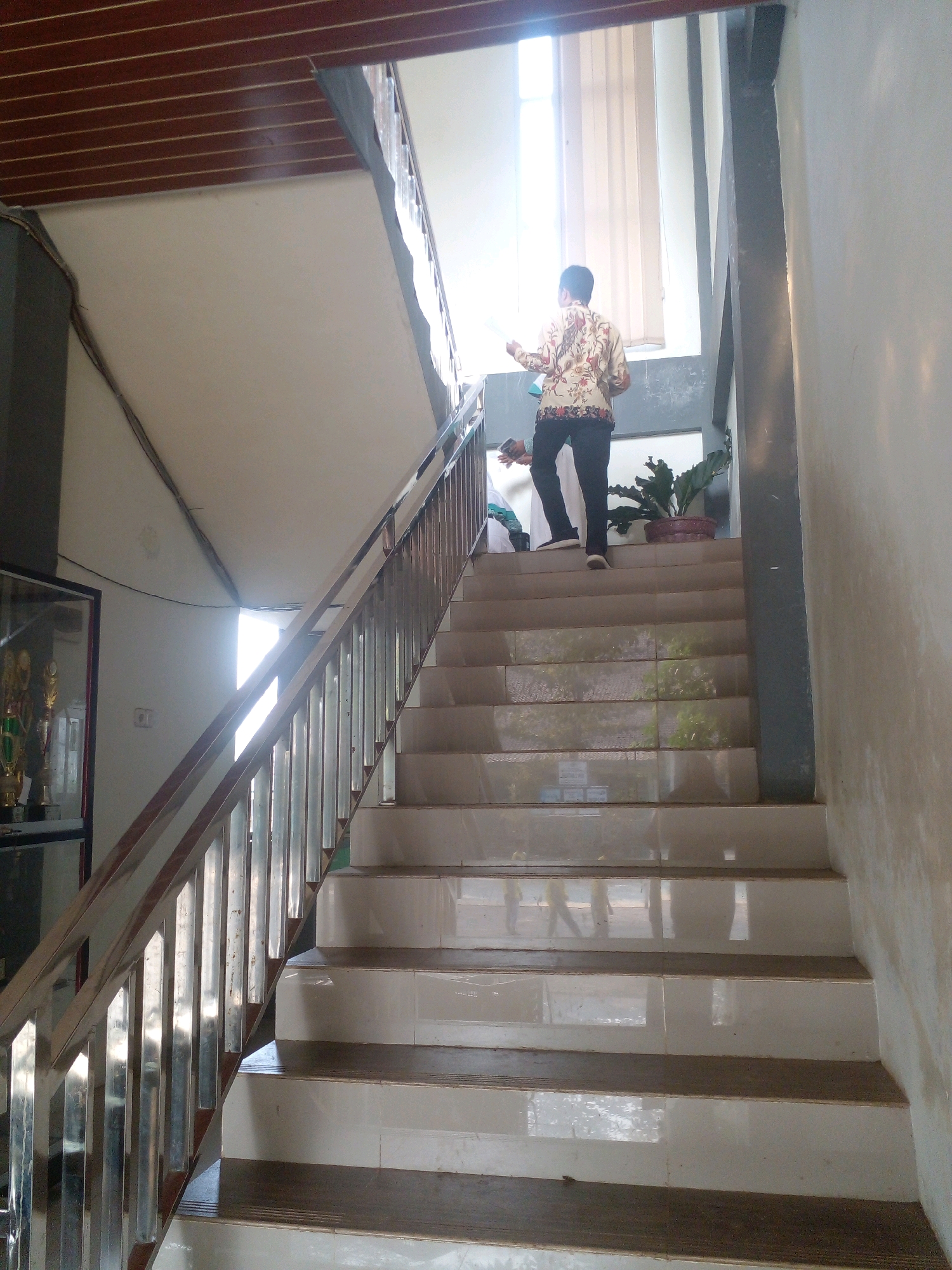 The image depicts a person walking up a tiled staircase with a metal handrail. The person is wearing a patterned shirt and dark pants. Another person is partially visible on a lower step. The staircase is located indoors, possibly in a building in Banyuputih, Indonesia, given the context. Sunlight streams in from a large window at the top of the stairs, creating bright highlights and shadows. A potted plant is visible on a landing. To the left of the staircase, a glass display case containing trophies is present. The overall environment suggests a public or institutional building.