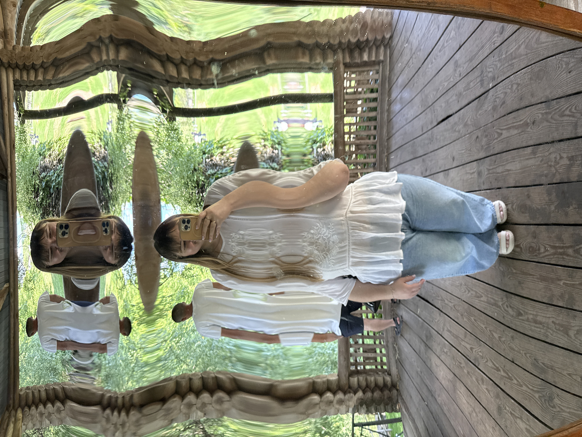 The image shows a distorted reflection in a wavy mirror, likely located outdoors in a rustic setting. In the foreground, a person is holding a phone and taking a selfie. They are wearing a white embroidered top and light blue jeans. To the left of the foreground person, another person wearing a long white garment and dark shorts is reflected. Further back, another reflection shows a person in a white t-shirt and dark pants. The background appears to be a lush green landscape with trees and foliage. The mirror is framed by rough wooden structures, suggesting a gazebo or a similar outdoor structure. The distortion in the mirror creates a warped and surreal effect on the reflections, making the scene appear fluid and elongated. The setting appears to be a park or garden area, possibly in Kotovo, Russia, given the description request, though there are no specific visual cues to confirm the location.