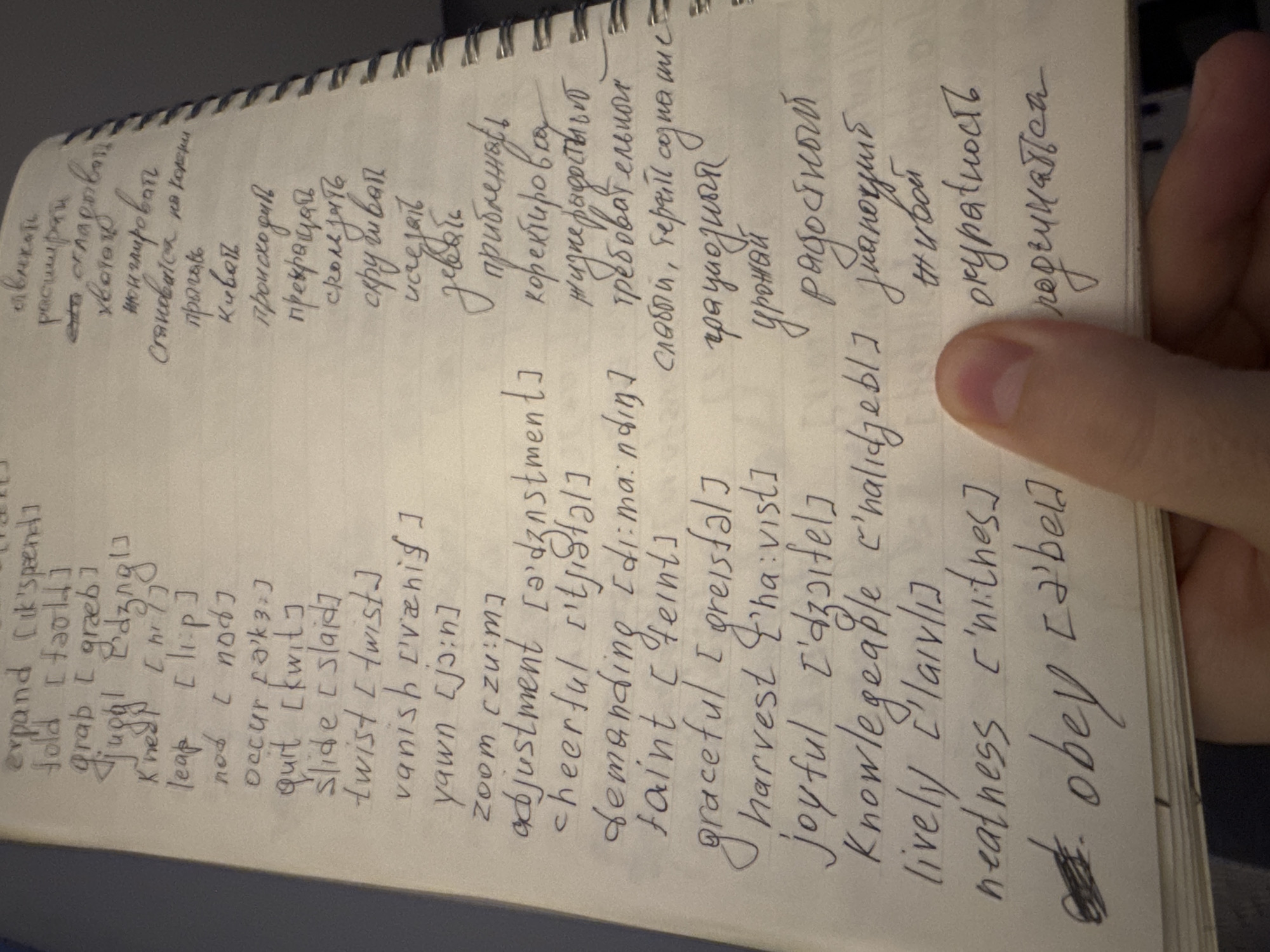 The image shows a handwritten notebook page with English words and their phonetic transcriptions on the left, and their Russian translations on the right. The notebook is spiral-bound. A hand with a visible thumbnail is holding open the notebook on the right side of the page.
