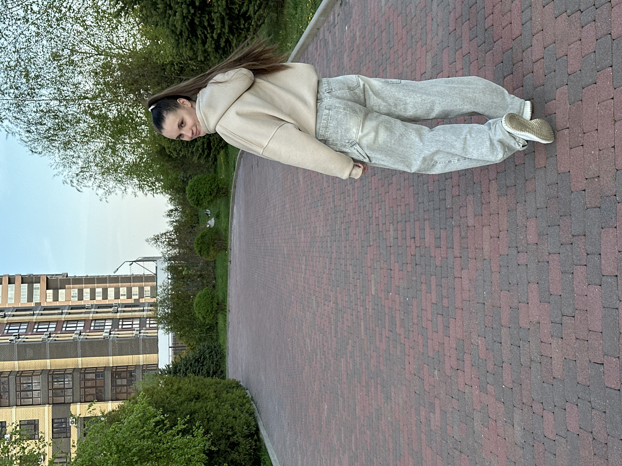 A young woman stands on a brick pathway in Rostov-on-Don, Russia. She wears a beige hoodie and light gray jeans, with matching beige sneakers. Her long brown hair is pulled back into a ponytail. The pathway curves and is bordered by green bushes and trees. In the background, a multi-story building with numerous windows can be seen. The sky is light blue.
